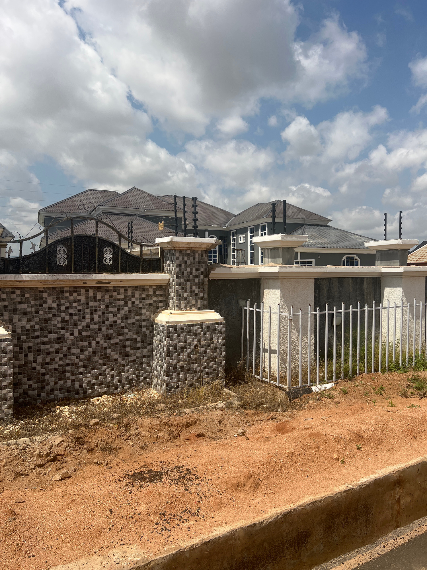 The image depicts a partially constructed modern building in Howayi, Nigeria. The building features a dark grey roof with decorative eaves, dark grey walls, and white trim around the windows and cornices. There is a white metal fence enclosing the property, with vertical bars and horizontal supports. The ground is covered with reddish-brown soil, and some dry vegetation is visible. In the foreground, there is a tiled wall with a mosaic pattern of dark and light grey square tiles, and a section of concrete steps. The sky is filled with scattered white clouds against a blue backdrop, suggesting it is daytime. There are no people visible in the image, and the scene appears to be a construction site or a recently completed residential property. The overall impression is one of ongoing development and a sunny, possibly warm, day.