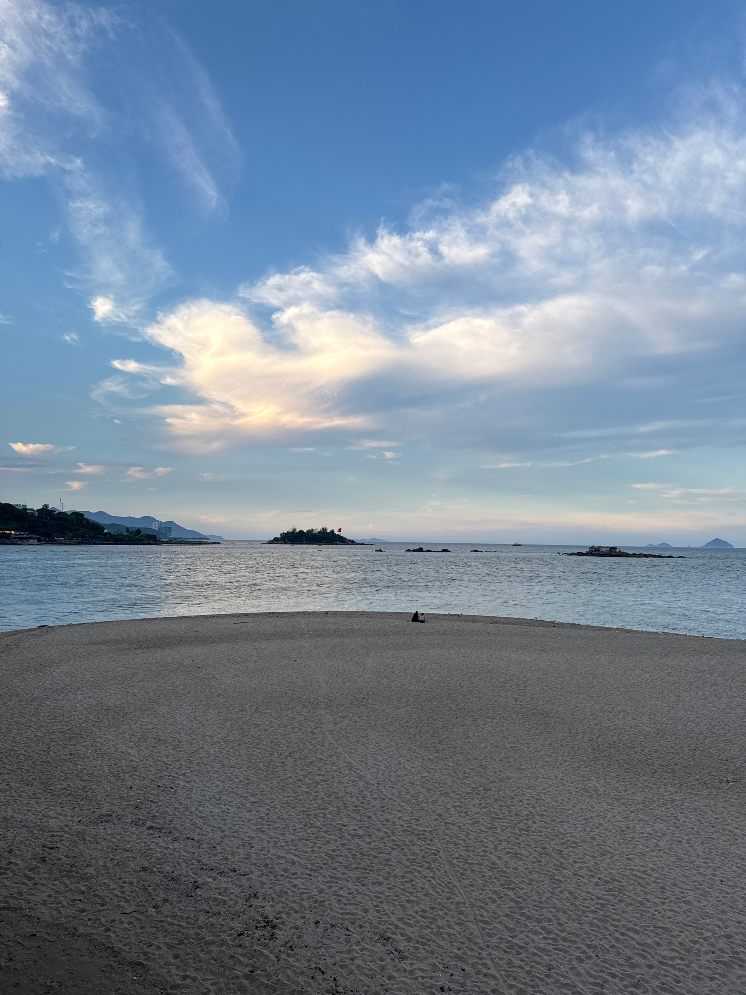 A beach scene is depicted with the camera rotated 90 degrees clockwise, making the horizon run vertically. The left side of the image is dominated by a vast blue sky with scattered white and golden clouds, illuminated by the soft light of either sunrise or sunset. The clouds are wispy and dynamic, suggesting movement.
The middle section of the image shows a calm, rippling body of water, likely the sea. A distant, narrow strip of land with trees and some buildings is visible along the horizon. The water reflects the colors of the sky, creating a soft, diffused light. A small island or rocky outcrop with structures on it can be seen further out in the water.
The right side of the image features a sandy beach. The sand has a textured surface with many small indentations, possibly from wind or previous waves. The sand appears damp near the water's edge and drier further inland. In the distance, on the beach, a single silhouetted figure can be seen, possibly a person sitting or standing. Another dark, indistinct shape further to the left might be another person or an object. The overall mood is serene and peaceful, evoking a sense of tranquility associated with a coastal landscape at dusk or dawn. The location is identified as Nha Trang Ward, Vietnam. There is no visible text in the image.