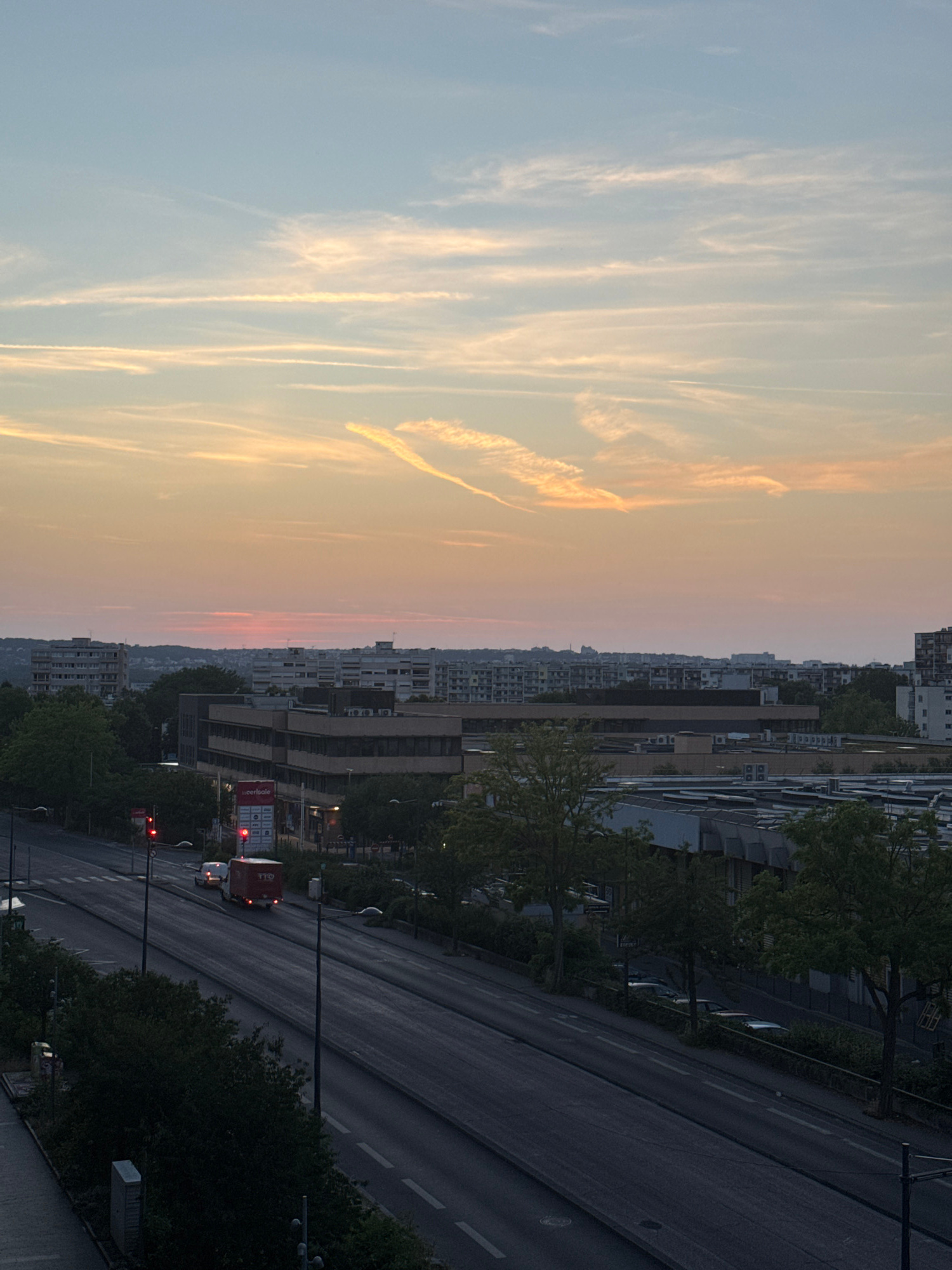 A medium shot captures a cityscape at dusk from an elevated perspective, looking towards the horizon where a soft, pastel sunset illuminates the sky with hues of orange, pink, and yellow. Wispy clouds streak across the pale blue expanse, some appearing as contrails from aircraft. In the midground, a cluster of multi-story buildings, characteristic of urban development, are visible. These buildings display a range of architectural styles, from the boxy, repetitive facades of apartment blocks to more modern, low-rise commercial structures with dark-tinted windows. Green trees, some with full foliage and others sparsely leafed, are interspersed between the buildings and along the edges of the roads, suggesting a degree of urban greenery.
In the foreground, a wide, multi-lane road runs diagonally from the lower left to the upper right of the frame. The road is paved and marked with white lines indicating lanes and pedestrian crossings. The scene is characterized by a relative lack of activity on the road. A single white van and a red truck with the markings "TID" are traveling away from the viewer on the right-hand lanes. Traffic lights are visible, with two displaying red signals. The overall lighting suggests late evening or early morning, with shadows beginning to lengthen. The environment appears to be a populated urban area, possibly on the outskirts of a city, given the mix of residential and commercial buildings and the presence of a major road. The sky dominates the upper half of the image, setting a tranquil yet potentially dynamic atmosphere.