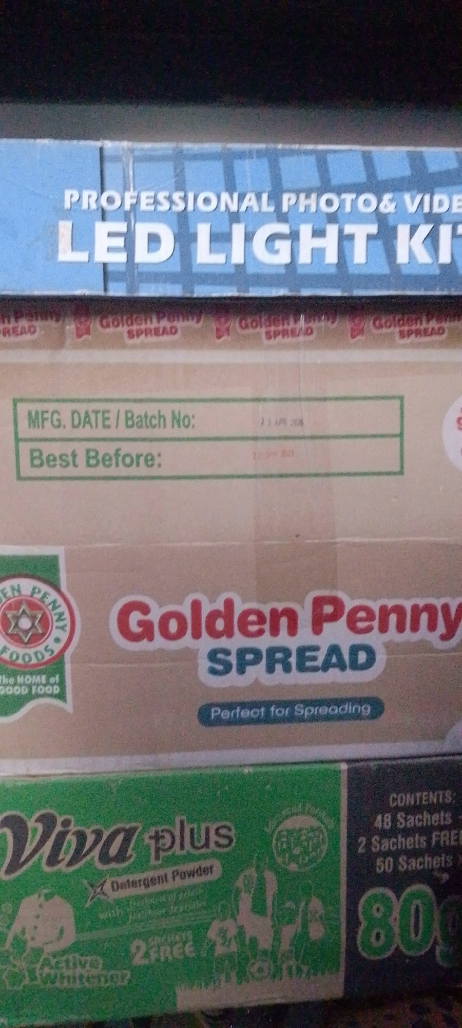 The image shows a stack of product boxes. The top box is blue and has white text that reads "PROFESSIONAL PHOTO & VIDEO LED LIGHT KIT". Below this, there are several boxes of "Golden Penny SPREAD". One of these boxes has a green and white label with fields for "MFG. DATE / Batch No:" and "Best Before:". The date provided is "23 APR 2020" and "23 JAN 2023". Another box in the stack is green and has the text "Viva plus Detergent Powder" and other graphics related to laundry. To the right of the "Viva plus" box, there is text that reads "CONTENTS: 48 Sachets, 2 Sachets FREE, 50 Sachets" and large numbers "80". The image is taken from a slightly elevated angle, looking down at the products. The lighting is dim, and the background is dark. The overall scene suggests a retail or storage environment.