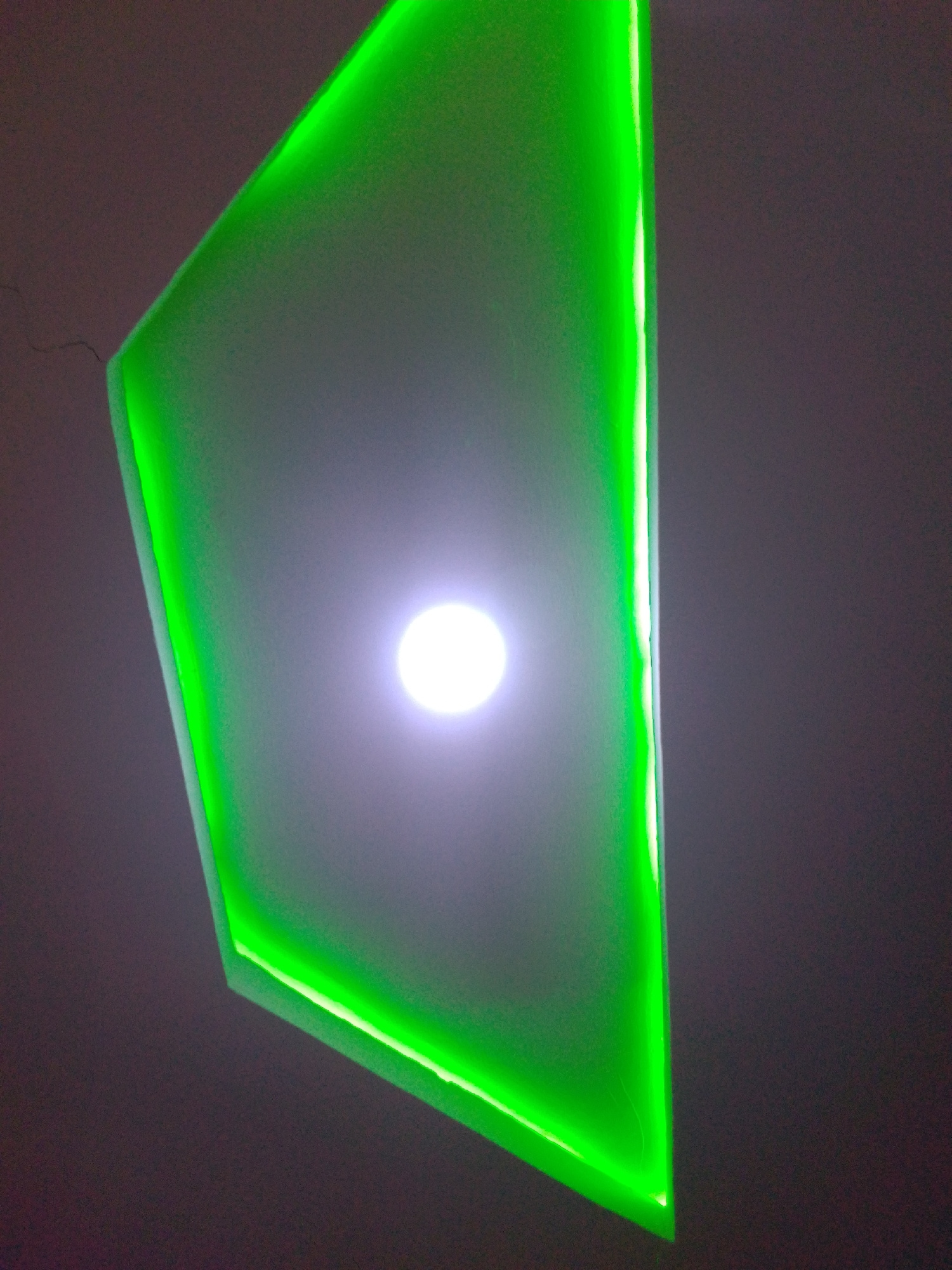 The image captures a modern ceiling design with integrated green LED lighting. The lighting is concentrated along the edges of a sharp, angular structure, which appears to be a recessed ceiling panel or a decorative element. A bright white light source is visible in the center of the panel, casting a glow that diffuses with the green lighting. The surrounding ceiling or wall is a dark, muted color, providing a stark contrast to the vibrant green illumination. The perspective suggests the photograph was taken from below, looking upwards at the ceiling. The scene appears to be an indoor environment, likely in a contemporary architectural setting. There are no people or discernible human activity visible in the image. The overall impression is one of modern, ambient lighting design. The location is stated to be in Howayi, Nigeria.