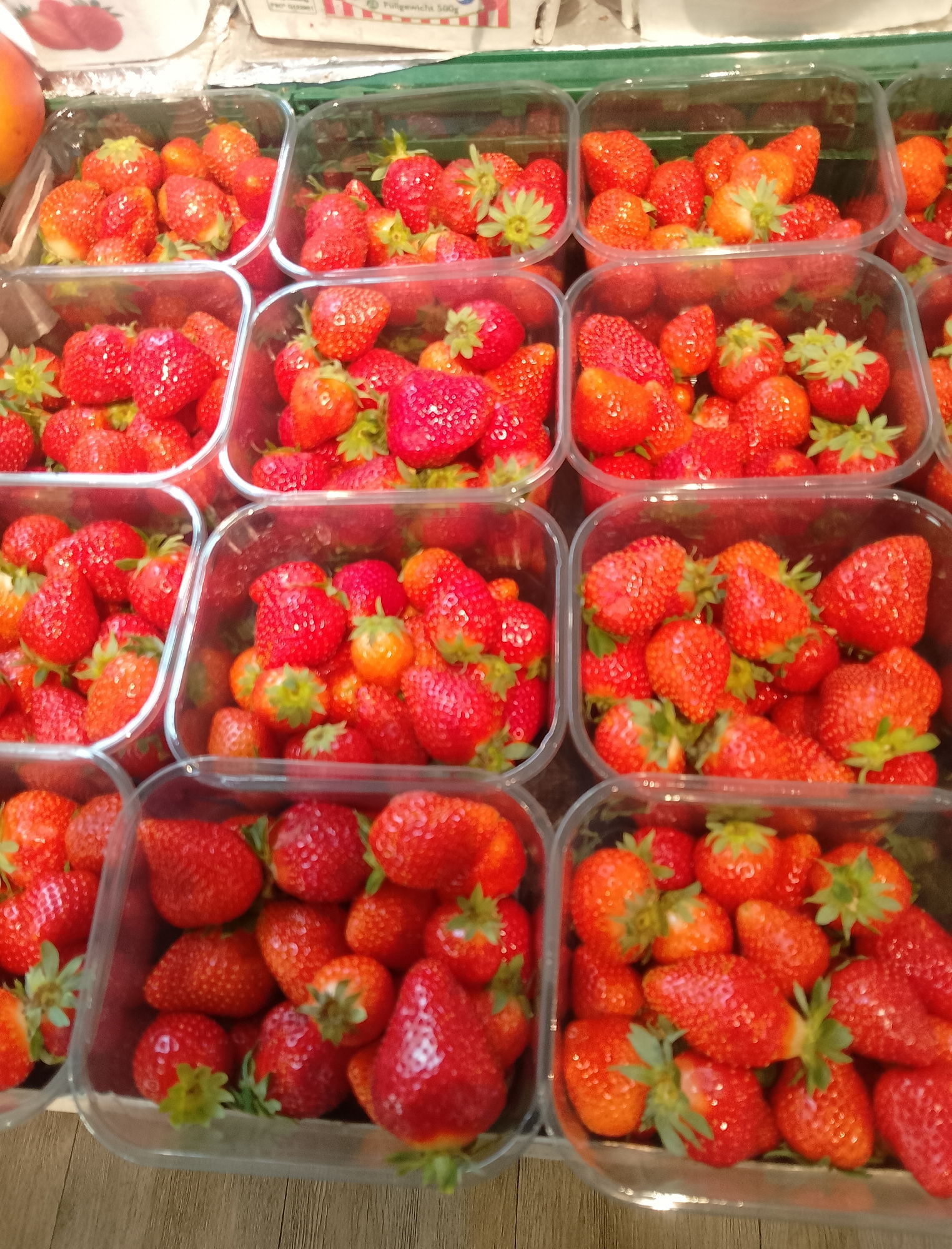 A top-down, close-up shot captures multiple clear plastic containers filled with fresh strawberries. The containers are arranged in rows, suggesting a display at a market or store. The strawberries vary in size and ripeness, with many appearing plump and a vibrant red. Some have visible green stems and leaves.
The lighting appears to be artificial, possibly from overhead store lights, as there are no shadows indicating natural sunlight. The background is blurred, but hints of wooden flooring or shelving are visible at the bottom of the frame. The overall scene is one of abundance and freshness, focusing entirely on the fruit. No people are visible. The strawberries are the sole subjects, presented in a clean and organized manner. The focus is on the fruit's texture and color.