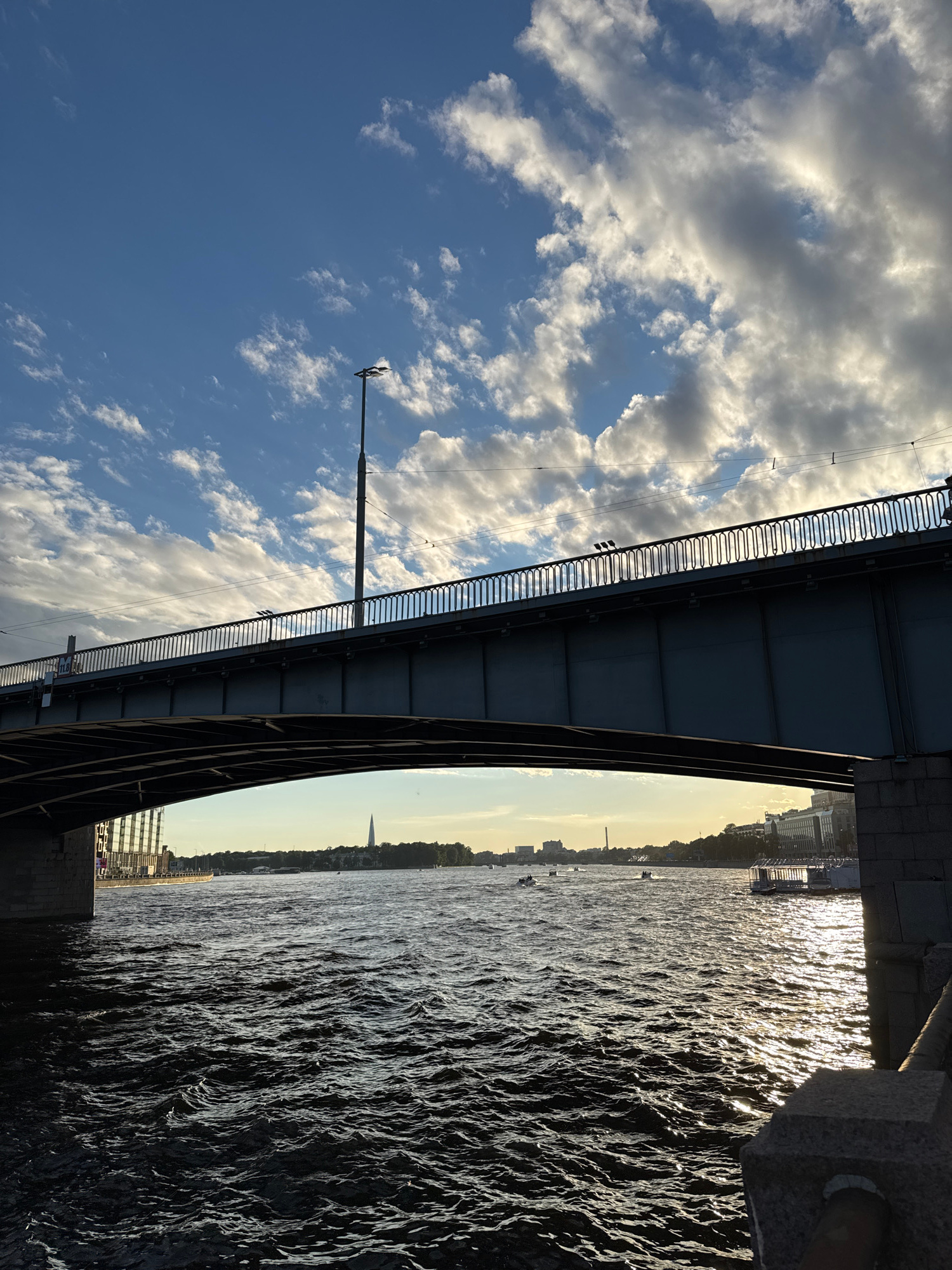 An eye-level, medium shot captures a wide, dark blue river flowing under a large, dark blue bridge. The bridge spans the entire width of the frame, with its underside casting a deep shadow on the water. A metal railing with decorative vertical bars runs along the top of the bridge, and a tall, dark lamppost stands on the bridge's walkway.
The water is choppy, with numerous small waves creating a textured surface that reflects the bright sunlight in streaks of gold and white. In the distance, across the river, a city skyline is visible. A tall, slender spire pierces the sky on the left side of the skyline. Several boats, appearing as small white shapes, are scattered across the water. The sky above is a vibrant blue, punctuated by scattered white clouds, some of which are illuminated by the sun, creating a dramatic contrast. The overall lighting suggests it is either early morning or late afternoon. The right side of the frame shows a stone embankment with rough-hewn blocks.