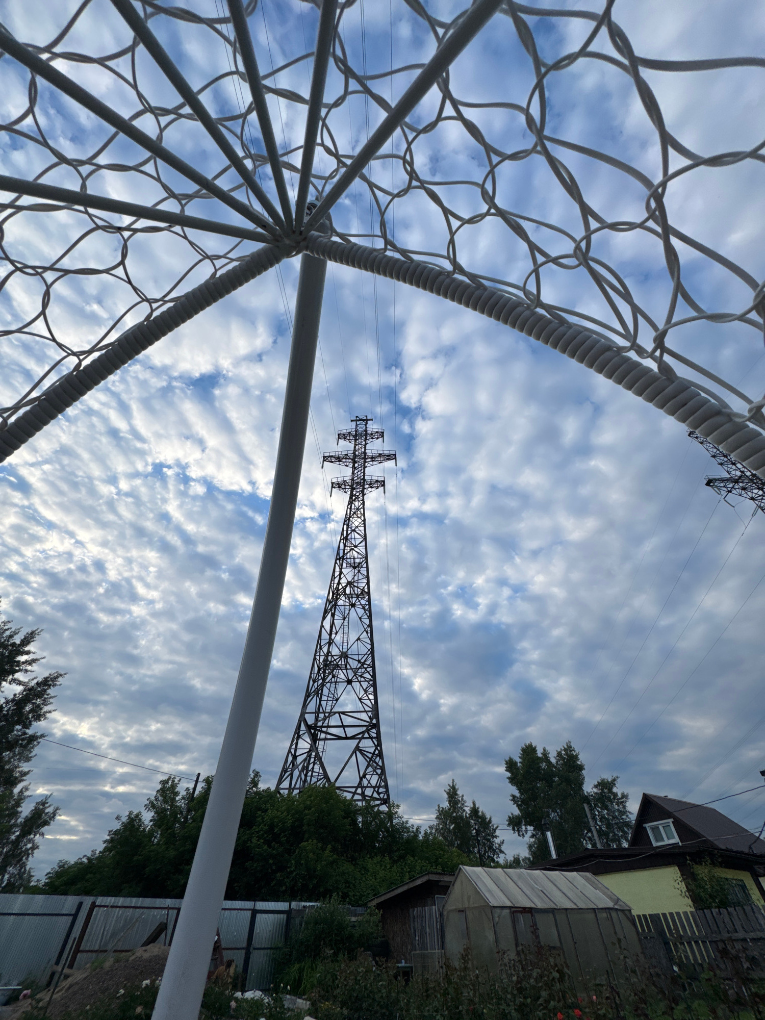 The image, captured in Chaikovsky, Russia, presents a low-angle perspective looking upwards towards a dramatic sky filled with cirrocumulus clouds. In the foreground, a portion of a white, decorative, possibly garden structure, with a lattice of intertwined loops, frames the view. A thick white pole, part of this structure, extends upwards. Dominating the mid-ground is a tall, latticed electrical pylon, its dark metal contrasting sharply with the sky. Several power lines extend from the pylon across the frame. Below the pylon and stretching across the lower part of the image are dense green trees and foliage. A corrugated metal fence runs along the bottom left. In the lower right, a yellow house with a dark roof and a small window is visible, along with a smaller, older wooden building and a greenhouse. The scene suggests an outdoor setting, possibly a backyard or garden area, with elements of both domestic life and industrial infrastructure.