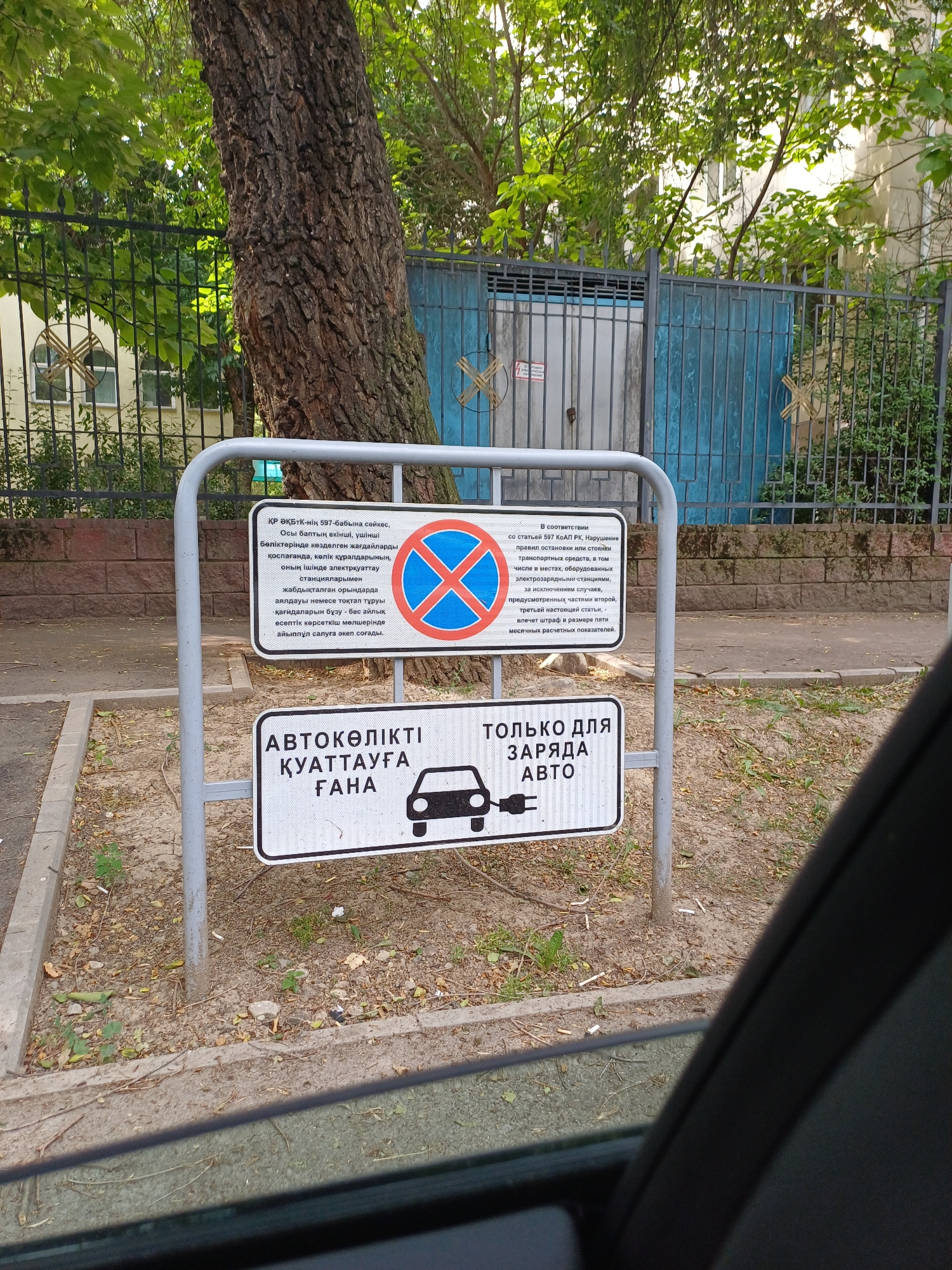 An outdoor scene captures a metal signpost supporting two rectangular signs. The top sign is white with text in Russian and a circular red and blue prohibition symbol indicating no stopping or parking. The bottom sign is also white, with text in Kazakh and Russian, and features a black silhouette of an electric car being charged. The signs are positioned at eye level, and behind them, a large tree trunk with rough bark is prominent. A dark metal fence with vertical bars extends across the background, partially obscuring a light-colored building. The ground around the signpost is covered with dry leaves and dirt, and a concrete curb runs along the edge of a paved area. The scene appears to be in Almaty, Kazakhstan.