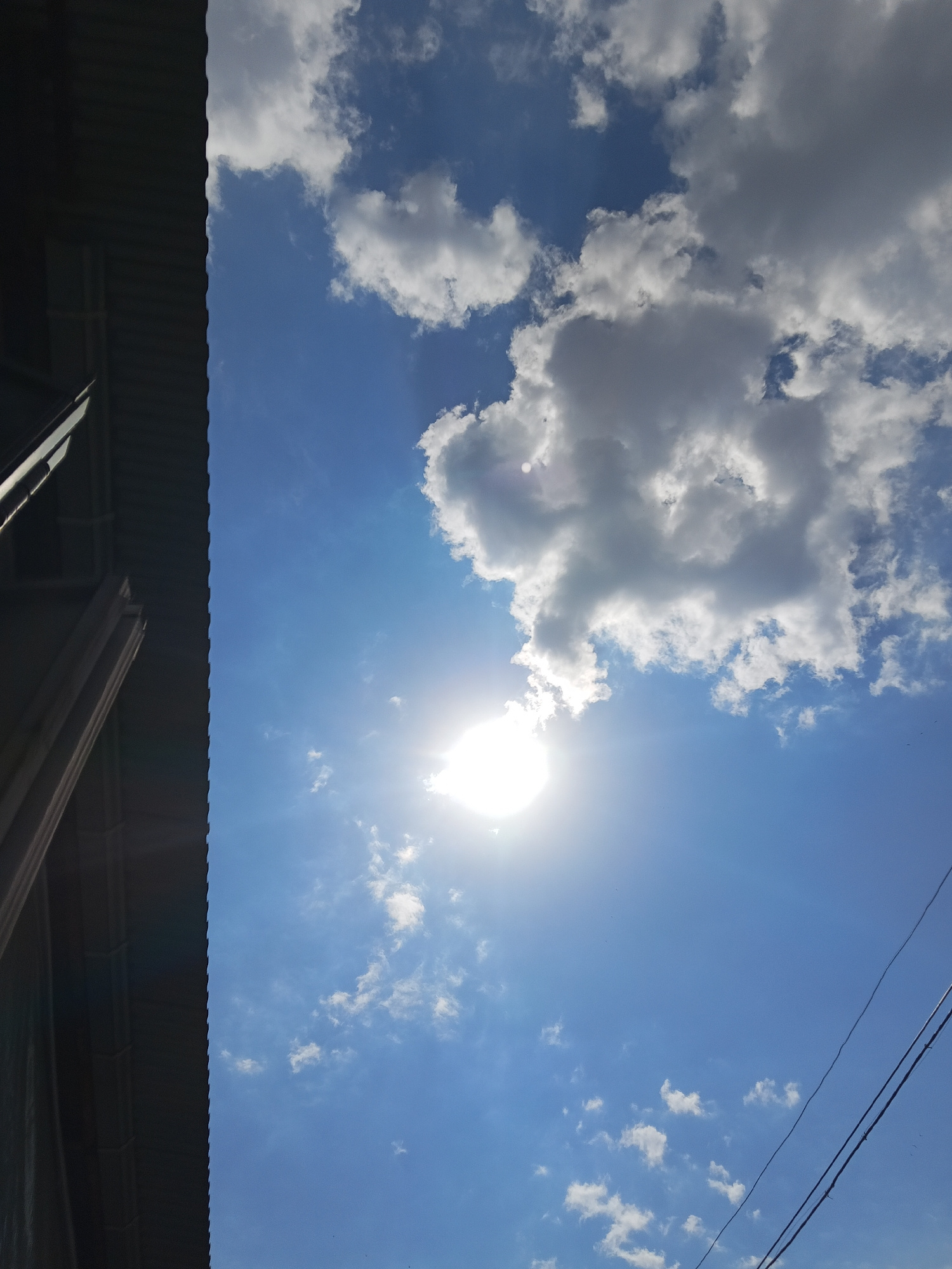 A low-angle, vertically oriented shot captures a bright sunny sky filled with fluffy white clouds and a prominent sunburst. To the left, the corrugated metal roof of a building angles upwards, partially obscuring the sky. The scene appears to be outdoors, likely during the daytime. There are no people or specific Yerevan landmarks visible to identify its precise location within the city. The environment suggests an urban setting due to the presence of the building and overhead power lines on the right side of the frame.