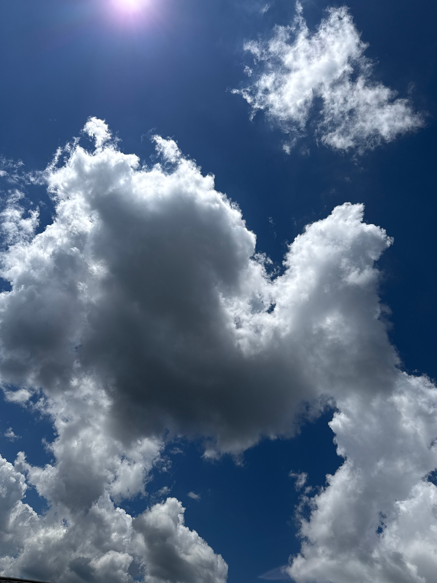 A clear, deep blue sky with scattered cumulus clouds is visible. Sunlight glints brightly in the upper left quadrant of the image. The clouds are predominantly white and fluffy, though some exhibit darker, shadowed undersides, suggesting varying depths and density. One large cloud formation dominates the center of the frame, appearing dense and voluminous. No people, structures, or specific landmarks are discernible in this sky-centric view. The image was captured in Hostomel, Ukraine.
