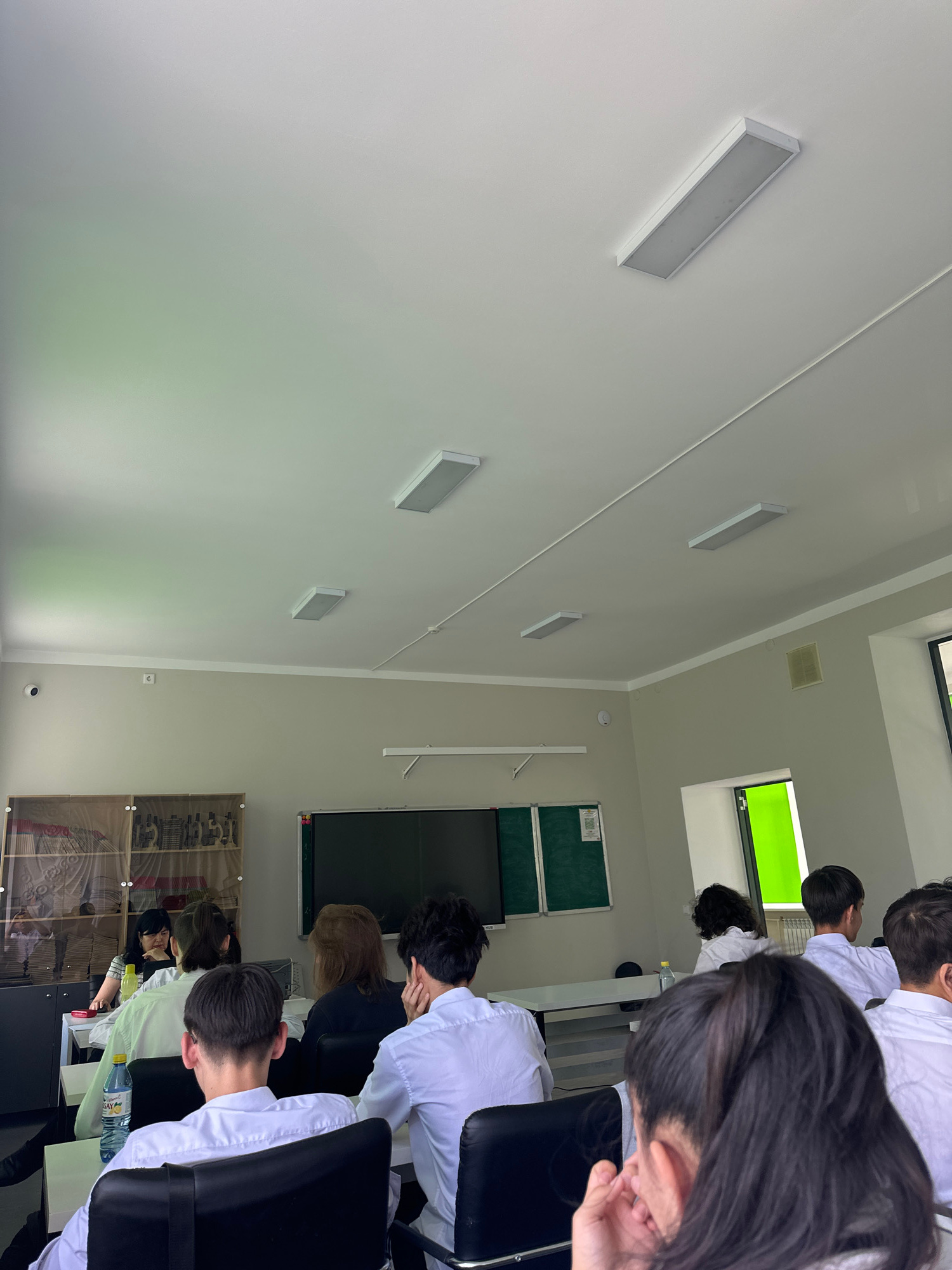 The image shows a classroom setting with several students seated at desks facing a teacher. The teacher is at the front of the room, near a whiteboard. There are fluorescent lights on the ceiling. The room appears to be a modern classroom with desks, chairs, and a whiteboard. The students are dressed in white shirts. There is a bottle of water on one of the desks.