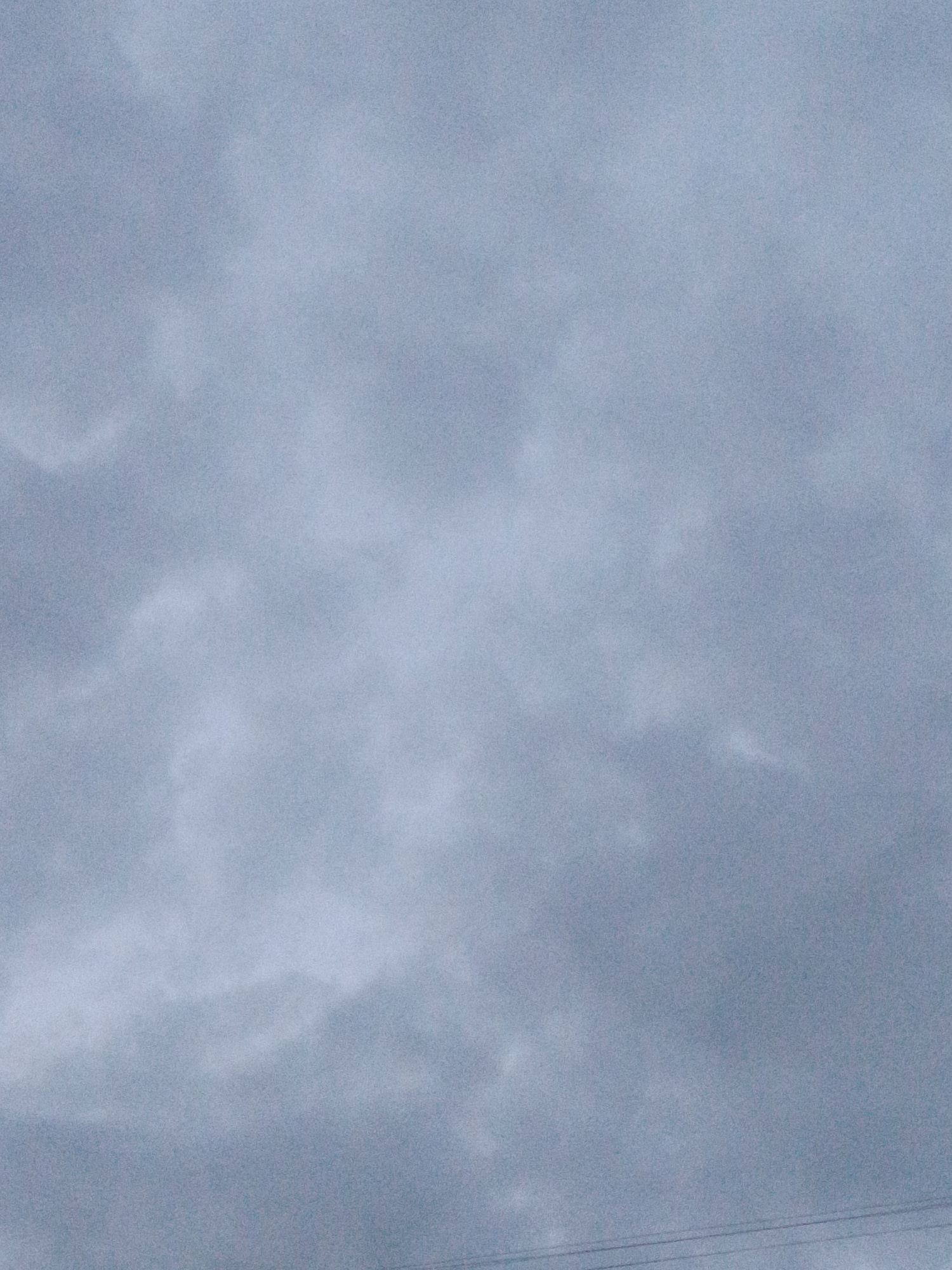 The image depicts an overcast sky with muted blue and gray tones. Wispy, diffused clouds are present, suggesting a cloudy or possibly rainy day. The overall mood is subdued and perhaps a bit melancholic due to the lack of direct sunlight and the uniform, gray lighting.
In the lower portion of the image, two thin, dark horizontal lines are visible. These appear to be electrical wires or cables, suggesting the presence of some form of infrastructure, such as power lines, in the scene. The wires are slightly out of focus, indicating they are in the foreground or mid-ground relative to the sky.
The image does not contain any people or discernible objects other than the electrical wires and the sky itself. The scene is abstract and focuses on the atmospheric conditions. Given the provided location context of Almaty, Kazakhstan, the cloudy sky is a common weather phenomenon in this region, particularly during certain seasons. The time of day is indeterminate as the lighting is diffused and lacks distinct shadows. There is no visible text in the image.