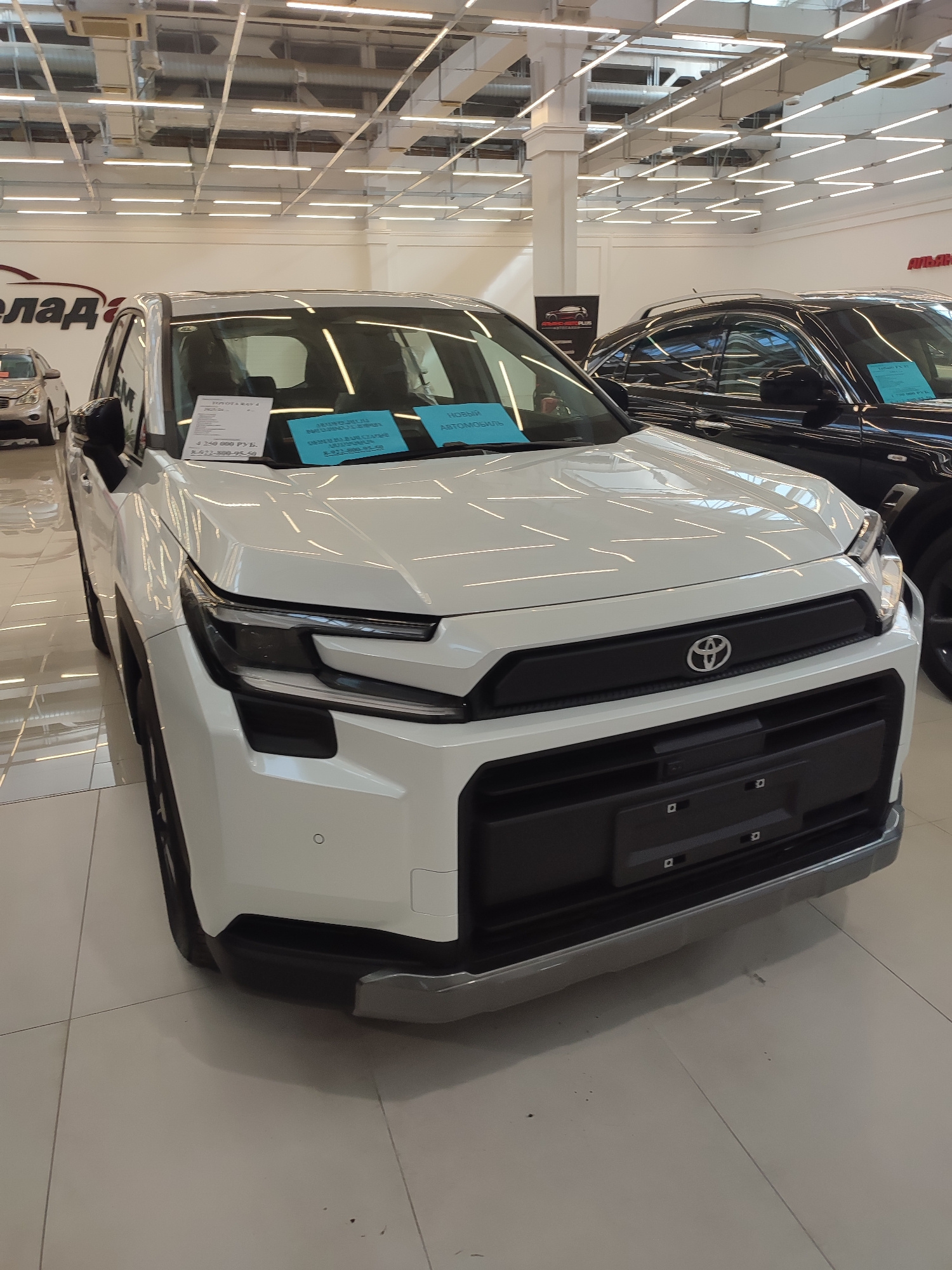 The image shows a white Toyota car parked inside a dealership showroom. The car is the main subject, positioned at a slight angle to the viewer, with its front fascia clearly visible. It has a prominent grille with the Toyota logo and distinctive headlights. There are two blue price tags on the windshield. In the background, another black car is visible, as well as some ceiling lights and retail shelving. The floor appears to be tiled. The dealership signage is partially visible, including the word "ЕЛАД" which is part of a Russian name, indicating a car dealership likely in Russia. Although the exact location is not explicitly stated in the image, the presence of Russian text on the signage and price tags suggests it is in Russia, and based on the provided context, is in the city of Orenburg, Russia. No people are visible in the frame.