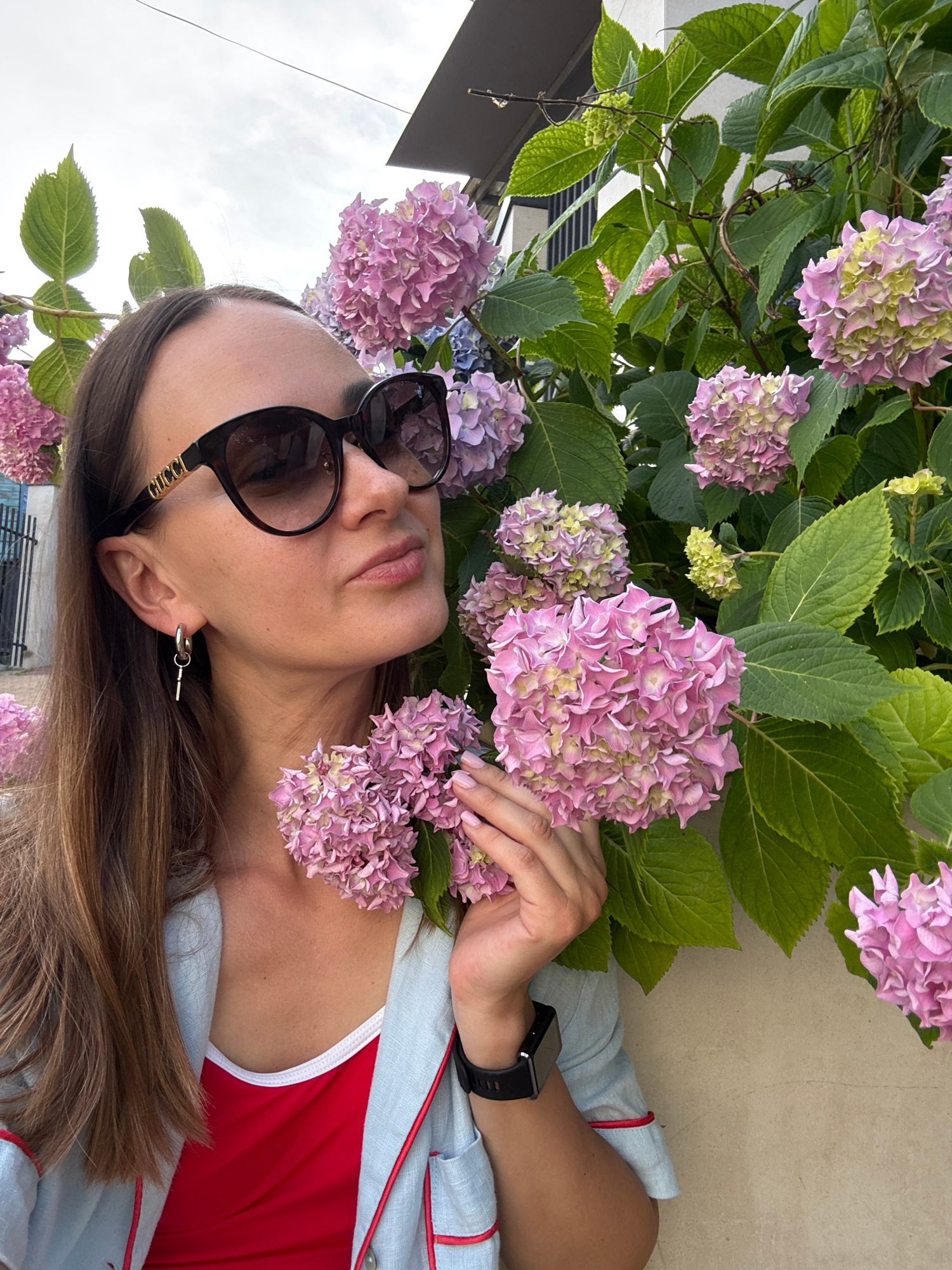 A woman, appearing to be in her late 20s or early 30s, with shoulder-length brown hair, is posing for a selfie in front of a wall and a dense bush of pink hydrangeas. She is wearing large, dark sunglasses with the "GUCCI" logo visible on the side, a silver earring in the shape of a key, a red tank top, and a light blue jacket with red trim. She is touching a cluster of hydrangeas with her right hand. The background shows a beige wall, a metal railing, and a building with a dark roof. The scene appears to be outdoors, likely in Sochi, Russia, given the floral environment and architecture. The sky is overcast.