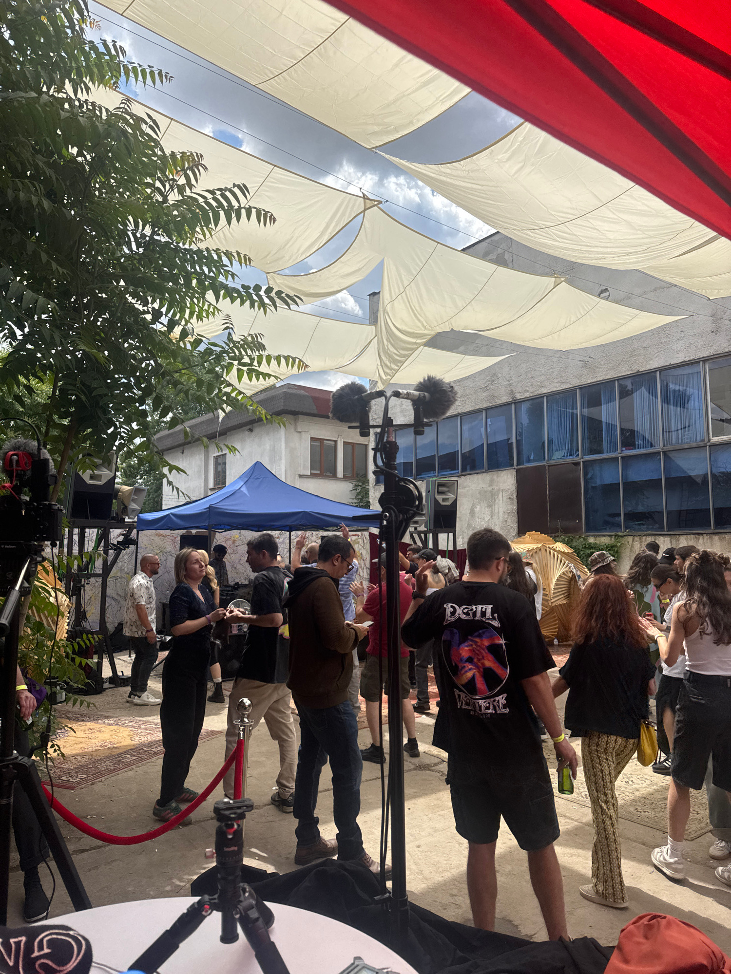 The image depicts an outdoor gathering in Sofia, Bulgaria, likely a festival or event. The scene is characterized by a casual atmosphere with a diverse group of people. Overhead, several white fabric canopies are stretched, creating a shaded area, with a red canopy visible at the top right. In the background, a building with large blue-tinted windows is present.
Several individuals are captured in various states of interaction. Some are standing and conversing, while others appear to be observing or moving through the space. A blue tent is set up, possibly serving as a vendor or information booth. There is a red rope barrier on the ground, suggesting an entrance or a designated area.
A prominent feature in the foreground is camera equipment, including tripods and microphones, indicating that the event is being filmed or recorded. Various people are dressed in casual attire, reflecting the relaxed nature of the gathering. The ground surface appears to be concrete, with a patterned rug visible in the middle ground. The presence of trees and foliage suggests an outdoor setting within a potentially urban environment. The overall impression is one of a vibrant social event with a touch of professional documentation.