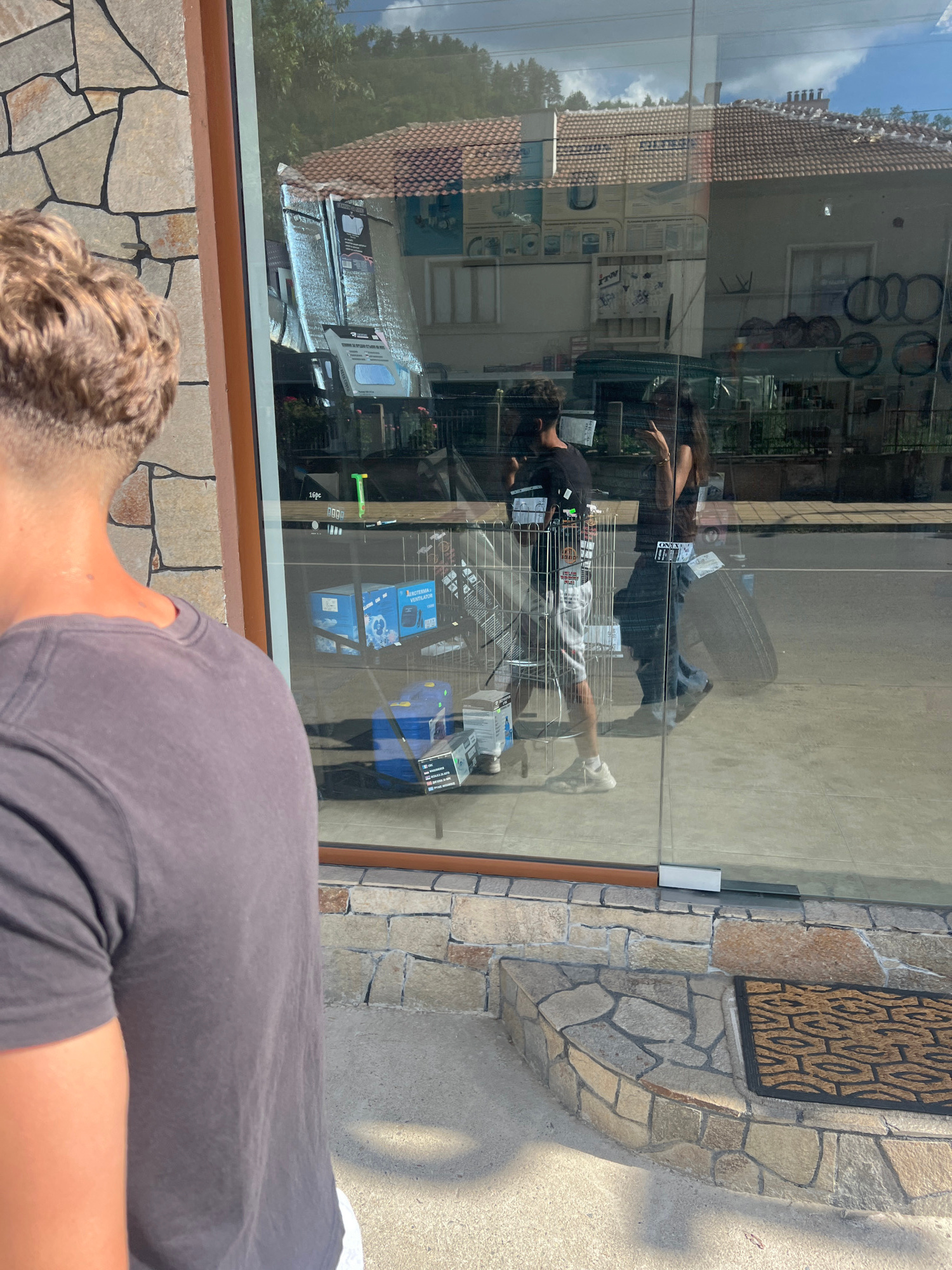 The image captures a reflection in a store window, showing two individuals and the interior of the store. In the foreground, the back of a person with curly hair and a grey t-shirt is visible. The reflection reveals a woman with long dark hair standing inside the store, wearing a black top and jeans, and a man in a black t-shirt and patterned shorts. The store interior appears to be a retail space, with shelves stocked with various items, including what look like boxes of electronics or appliances. The reflection also shows a tire inside the store.
Outside the window, the reflection captures a building with a tiled roof and windows, suggesting an urban or semi-urban setting. The ground outside is paved with concrete, and a stone facade is visible on the right side of the image, featuring a decorative mat. The lighting suggests it is daytime, with sunlight casting shadows. The overall mood appears casual and everyday.
Visible text includes labels on some of the products inside the store, though they are difficult to read clearly due to the reflection and image resolution. Some of the text appears to be in a Latin-based alphabet, possibly Bulgarian, given the location context of Zlatograd, Bulgaria. The reflection of the sky with clouds indicates a clear day.