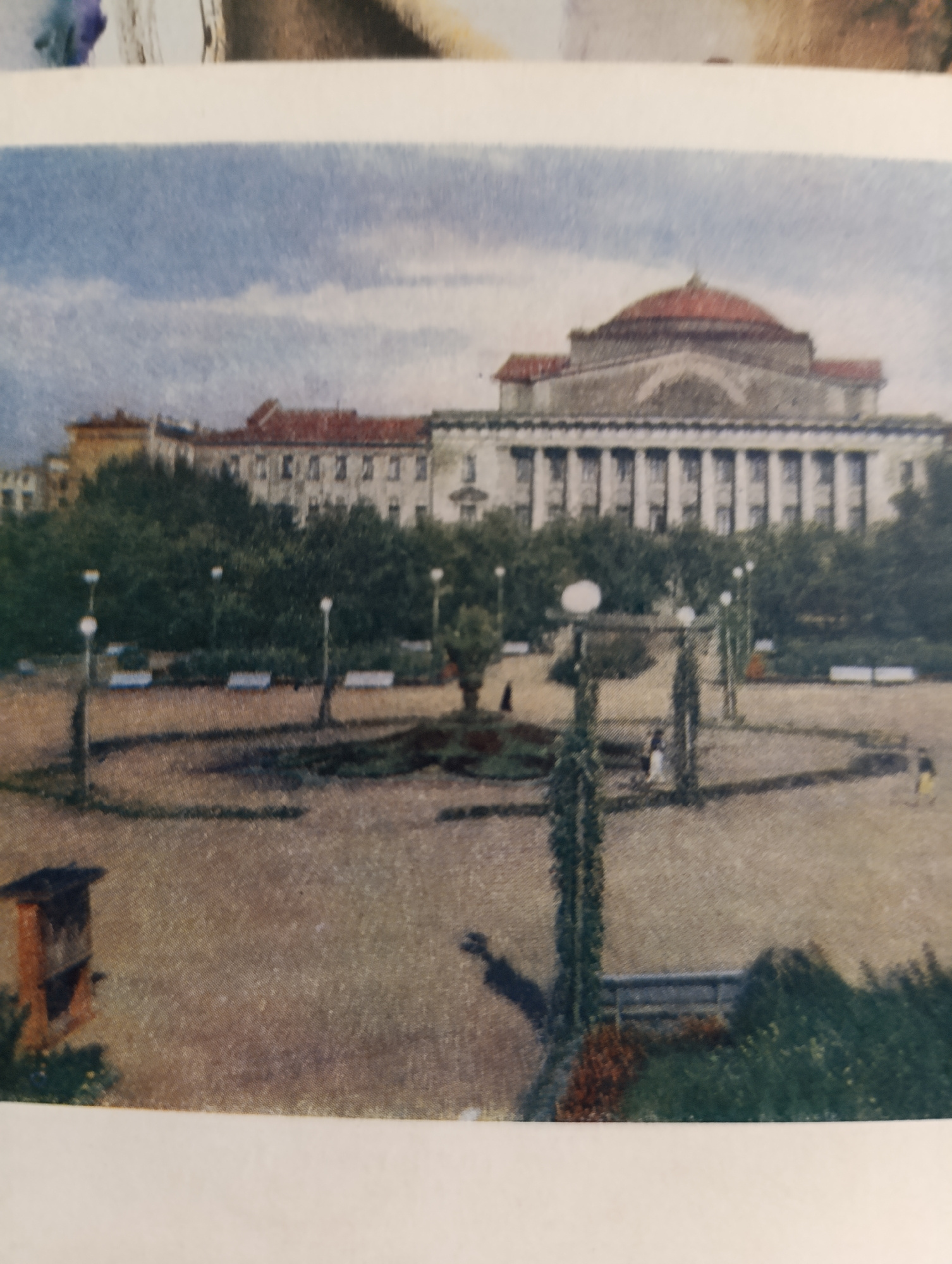 This is a color photograph of an outdoor public space in front of a large, neoclassical building with a prominent dome. The building has a long facade supported by a colonnade. In the foreground, there is a paved area with several lampposts and benches. Trees and bushes are visible, some of which are climbing structures. A few indistinct figures are scattered throughout the scene, suggesting a park or plaza. The sky above is cloudy. The overall impression is of a formal urban landscape.