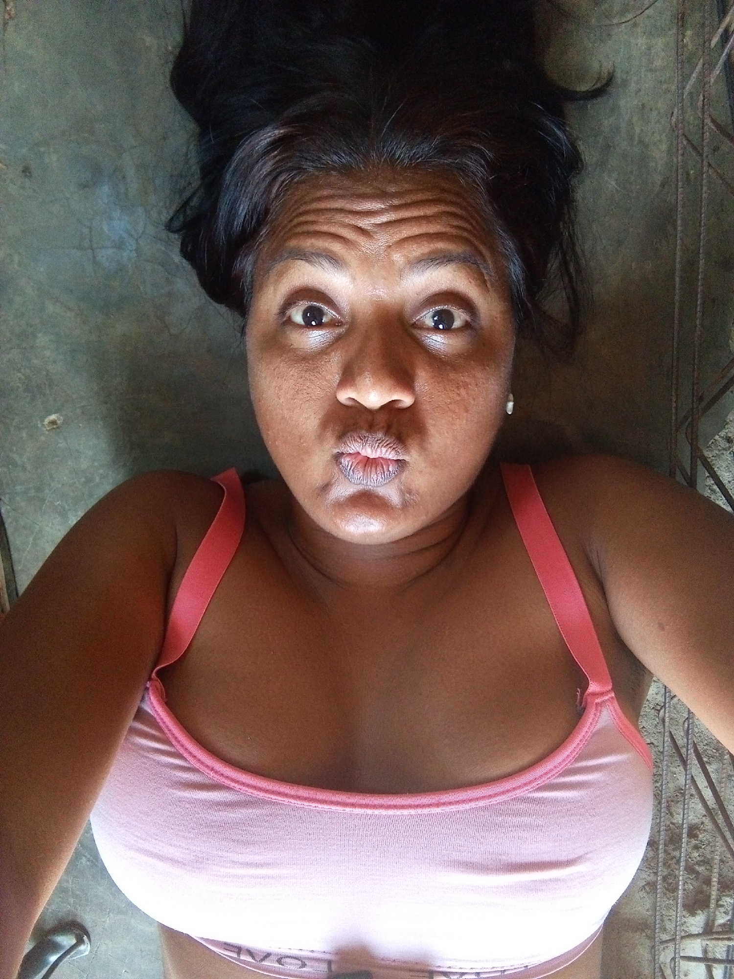 A high-angle, medium shot shows a woman with her head tilted back, lying on a textured concrete surface. Her dark hair is spread out behind her. She is wearing a light pink tank top or camisole with pink straps. Her face is positioned centrally, with her lips puckered, as if blowing a kiss. The background consists of the concrete floor and a blurred area to the right that appears to be a metal grate or fence.