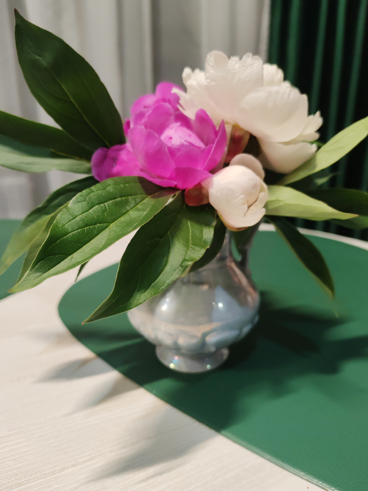 A still life image features a small, iridescent vase holding a bouquet of pink and white peonies with green leaves. The vase is placed on a surface divided into a green section and a white section. In the background, sheer curtains are visible. The image does not contain information about Komsomolsk-on-Amur, Russia.