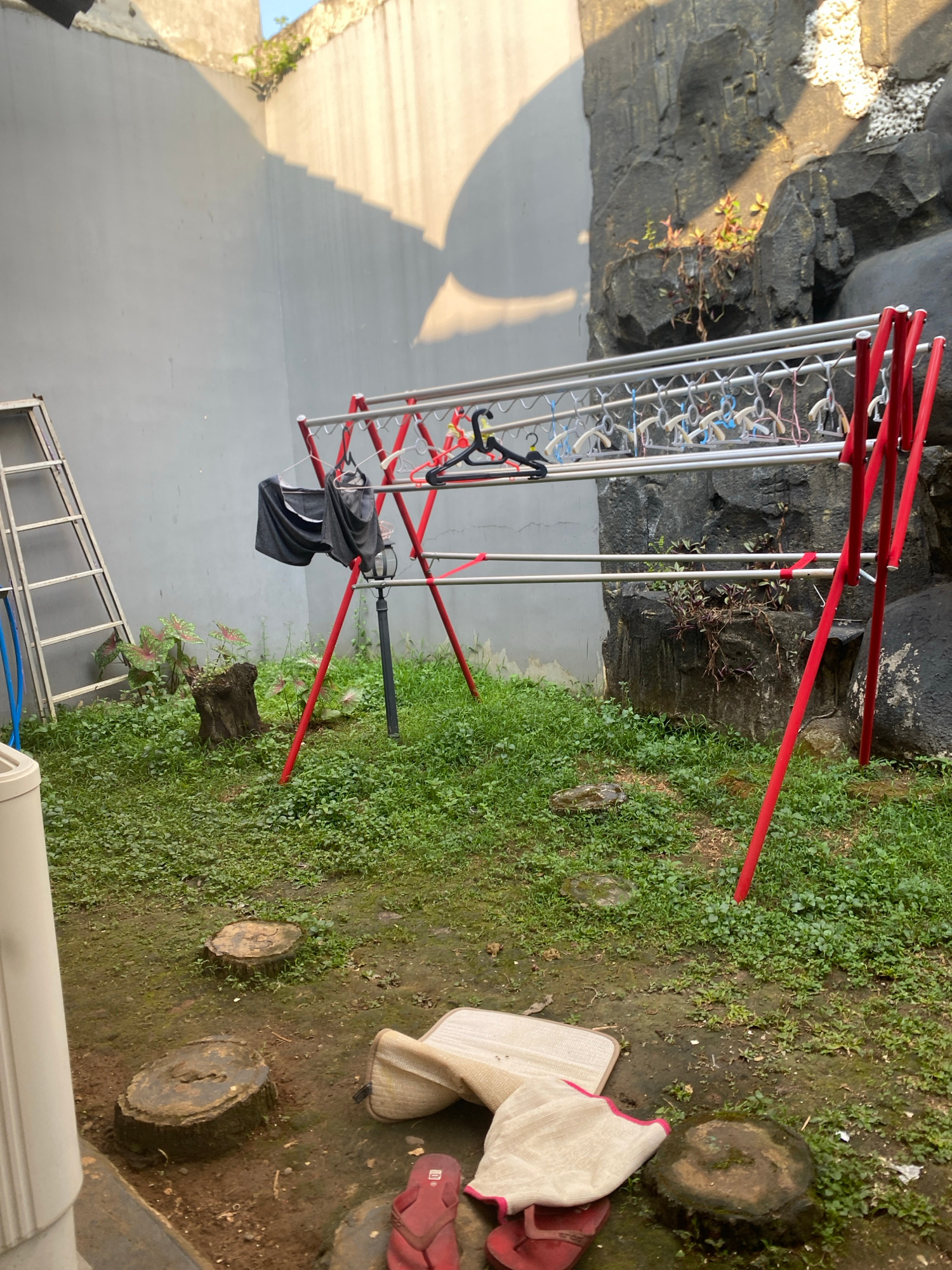 In Dukuh, Indonesia, a backyard scene unfolds under a bright sky casting sharp shadows on a gray wall. A red and silver clothes drying rack is prominent, holding several black and gray garments on hangers, along with numerous empty plastic hangers. To the left, a silver ladder leans against the wall, its blue hose coiled nearby. The ground is covered in lush green grass and scattered tree stumps, with a few plants growing near a weathered wooden stump. In the background, a rough, gray rock formation rises, dotted with small green plants. In the foreground, a rolled-up beige mat and a pair of red flip-flops lie on the ground, near a white appliance on the left.