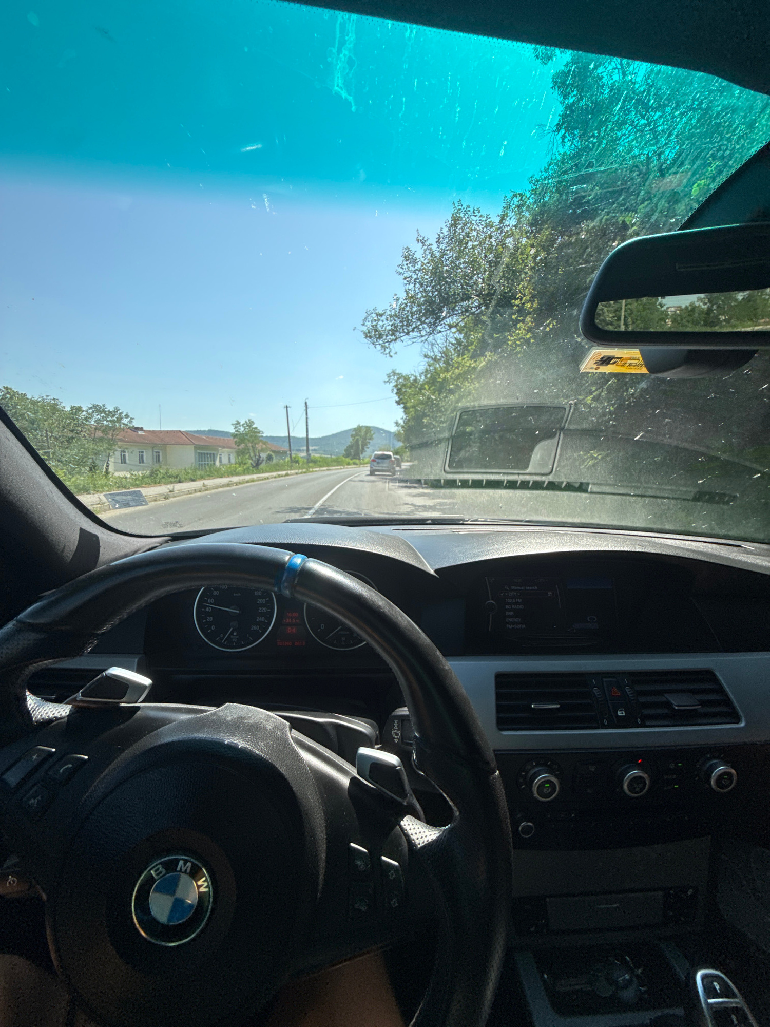 The image is a first-person perspective from inside a car, likely a BMW, driving on a road. The steering wheel and dashboard are visible in the foreground. The road is asphalt and appears to be in a rural or semi-rural area, with trees and vegetation on the sides. In the distance, there are houses and hills under a clear blue sky. A car is visible ahead on the road. The scene captures a moment of travel. The location is identified as being in the city of Dryanovo, Bulgaria.