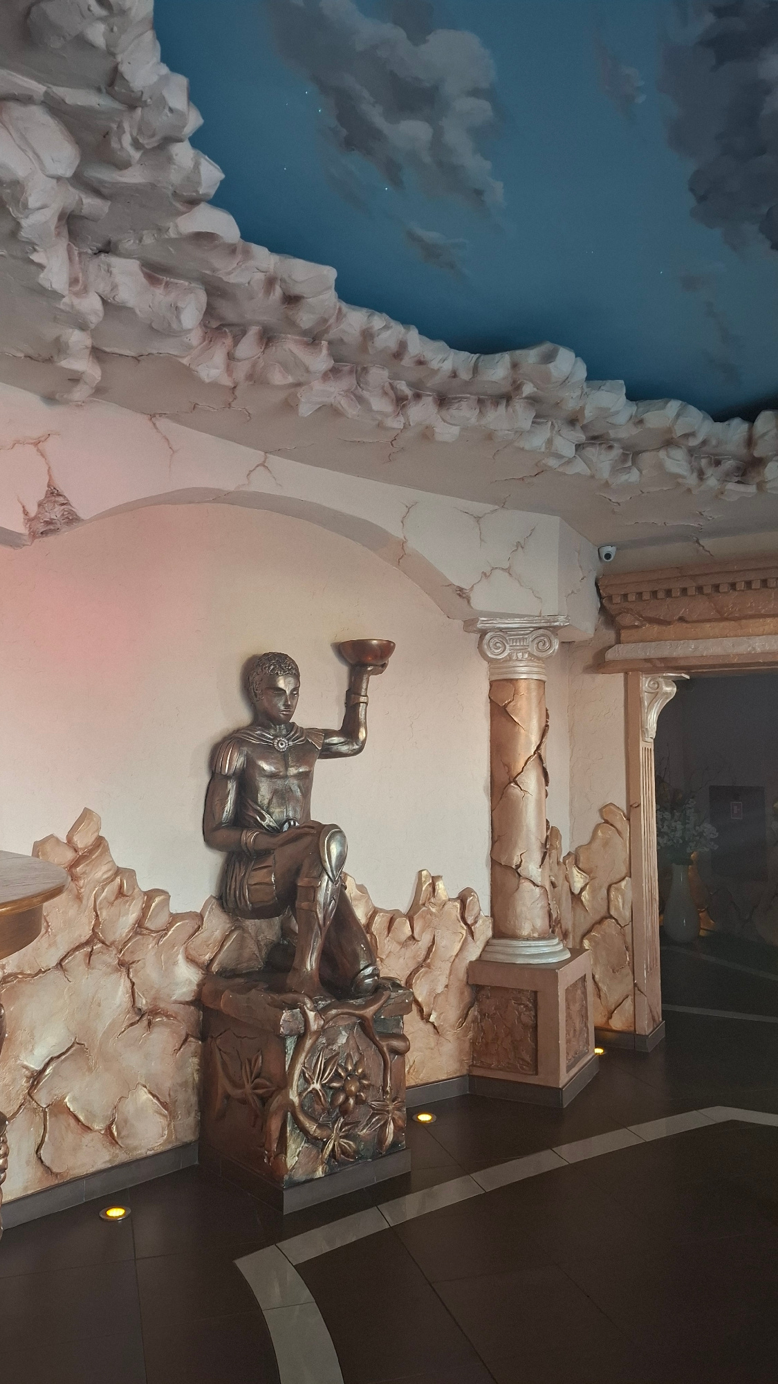 The image depicts an interior scene featuring a decorative ceiling painted with a blue sky and clouds, accented by textured cloud-like structures along the edges. A central focus is a bronze-colored statue of a young man holding a bowl aloft. The statue is seated on a decorative base adorned with floral motifs. The background includes a fluted column with a Corinthian capital, and a wall with cracked plaster detailing. The floor is tiled in dark brown with a lighter inlaid pattern. The overall ambiance suggests an elaborate, possibly themed, interior space. The city of Pokrzywnica, Poland, is mentioned in relation to the location, but no specific landmark or establishment is identifiable from the image itself.