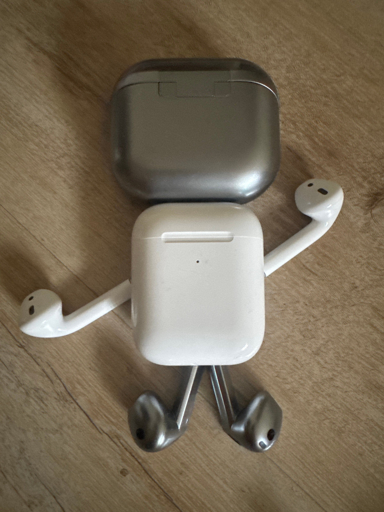 Two charging cases for wireless earbuds, one silver and one white, are placed side-by-side on a light brown wooden surface. The silver case is on the left, and the white case is on the right. Three wireless earbuds are visible. Two white earbuds are connected to the white charging case, extending outwards. One white earbud is on the left, pointing upwards, and another white earbud is on the bottom, pointing downwards. A third earbud, which appears to be silver, is emerging from the right side of the white charging case, pointing to the right. The wooden surface has a visible grain pattern and appears to be indoors. There is no indication of the time of day, weather, or specific location beyond the general context. No text is visible in the image.
