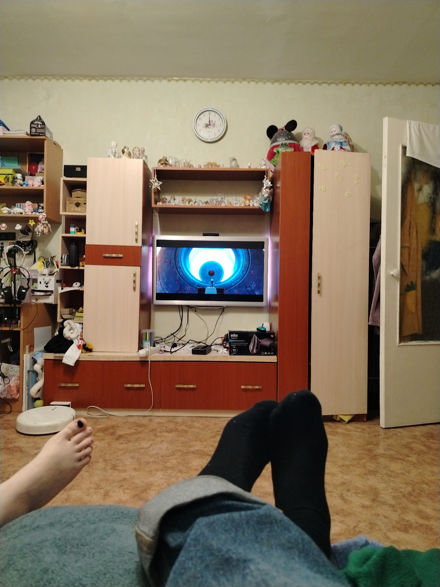 The image depicts a cozy living room scene, likely in Krasnoyarsk, Russia. The primary subjects are a pair of feet, one bare with black nail polish, and the other wearing a black sock, resting on a textured blue blanket. The feet belong to the person capturing the image, suggesting a relaxed, informal moment.
The setting is a room filled with furniture and decor. Dominating the center is a large entertainment unit with a television displaying a colorful animated scene. Adjacent to the television are various shelves and cabinets adorned with numerous small figurines, including what appear to be Santa Claus figures and small dolls, hinting at a festive or collectible theme. A white round clock with Roman numerals is visible on the wall above the entertainment unit, suggesting it is daytime. To the right, a tall, light-colored cabinet stands next to a partially open door, revealing clothing. On the floor to the left of the entertainment unit, a white robotic vacuum cleaner is positioned.
The overall atmosphere is one of comfort and leisure, with the focus on relaxation and entertainment. The warm lighting suggests it might be evening or a dimly lit day, enhancing the cozy feel. The presence of numerous decorative items adds a personal touch to the space. No specific weather is indicated, but the indoor setting implies a need for comfort away from the elements. The image conveys a sense of a quiet evening spent at home.