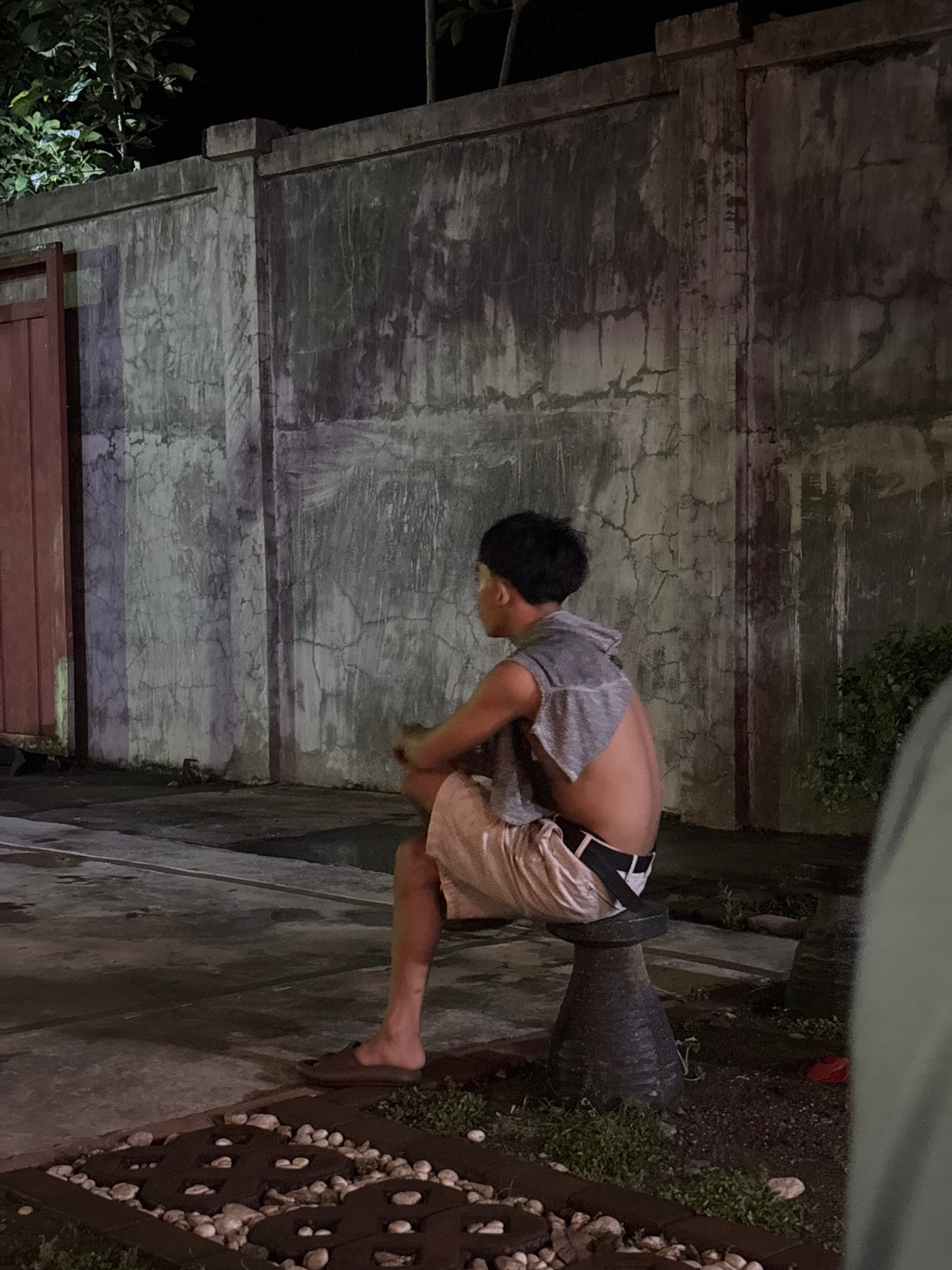 A young man with dark hair, wearing a grey sleeveless hoodie and light-colored shorts, sits on a dark, tapered stool facing left, in front of a textured concrete wall. The man's back is partially visible, and he appears to be looking away from the camera. The wall has vertical streaks and cracks, illuminated by light that casts shadows. To the left of the man, a portion of a reddish-brown door is visible. The ground in front of the wall is paved, with a decorative section filled with small white stones and dark geometric shapes. The environment suggests an outdoor area at night. The scene is located in Tagum City, Philippines.