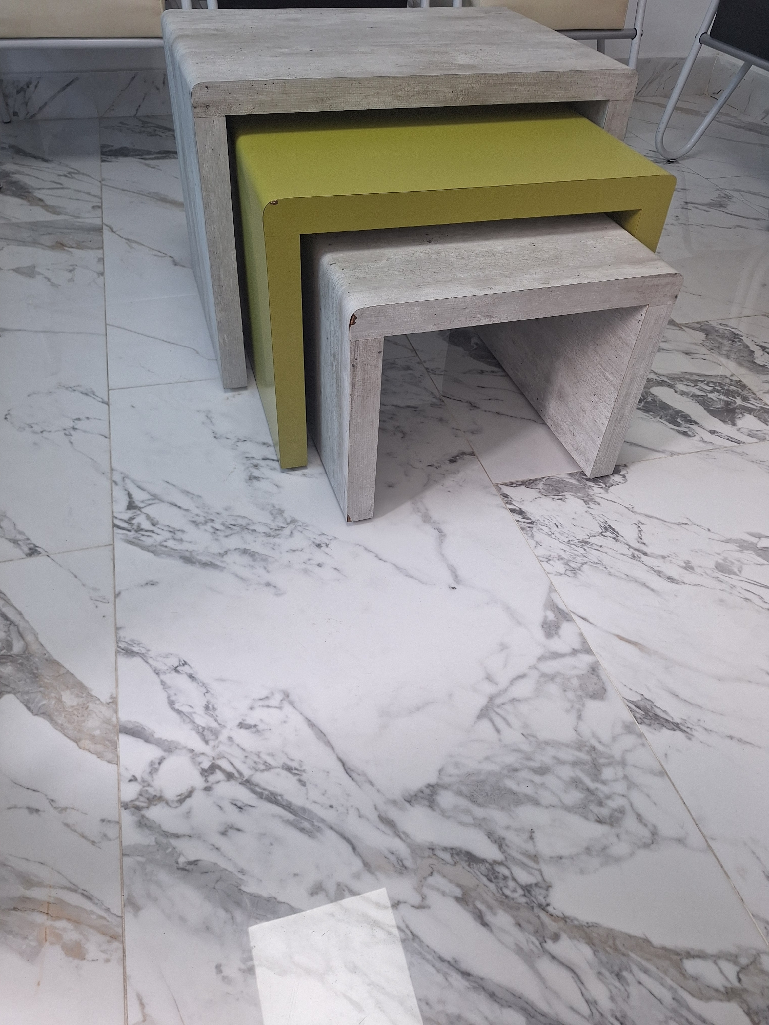 In Casablanca, Morocco, three nesting tables are arranged on a marble-patterned floor. The largest table, made of a light-colored wood or concrete-like material, is at the back. Inside it, a medium-sized table with a vibrant yellow-green surface is nested. The smallest table, also made of the light-colored material, is nestled inside the yellow-green one. The scene is captured from a slightly elevated angle, focusing on the tables and the floor.