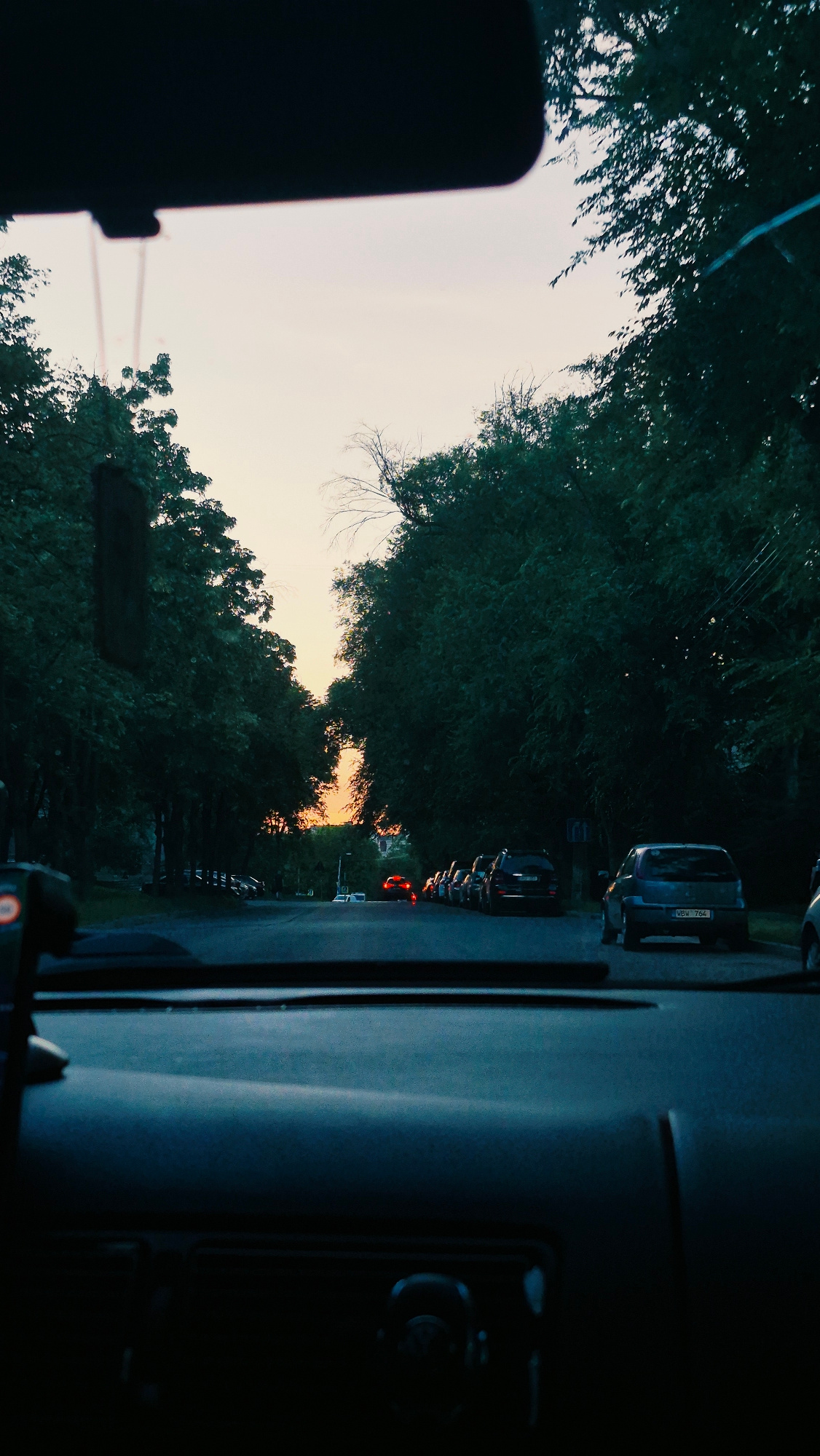 The image is a forward-facing shot from inside a car, looking out through the windshield at a tree-lined street during dusk or dawn. The sky displays a soft gradient of light orange and pale yellow, indicating either sunrise or sunset. On either side of the asphalt road, dense green trees create a canopy effect, with some branches extending over the road. A line of parked cars is visible along the right side of the street, and further down the road, a few cars are in motion, with their brake lights illuminated, suggesting a moderate amount of traffic. The interior of the car is dimly lit, with the dashboard and windshield frame visible in the foreground. One of the parked cars on the right has a license plate that reads "VBW 764". The overall atmosphere is peaceful and suburban. Based on the license plate format (three letters followed by three numbers) and common vehicle registration practices in Moldova, this scene is likely located in Chișinău, Moldova.