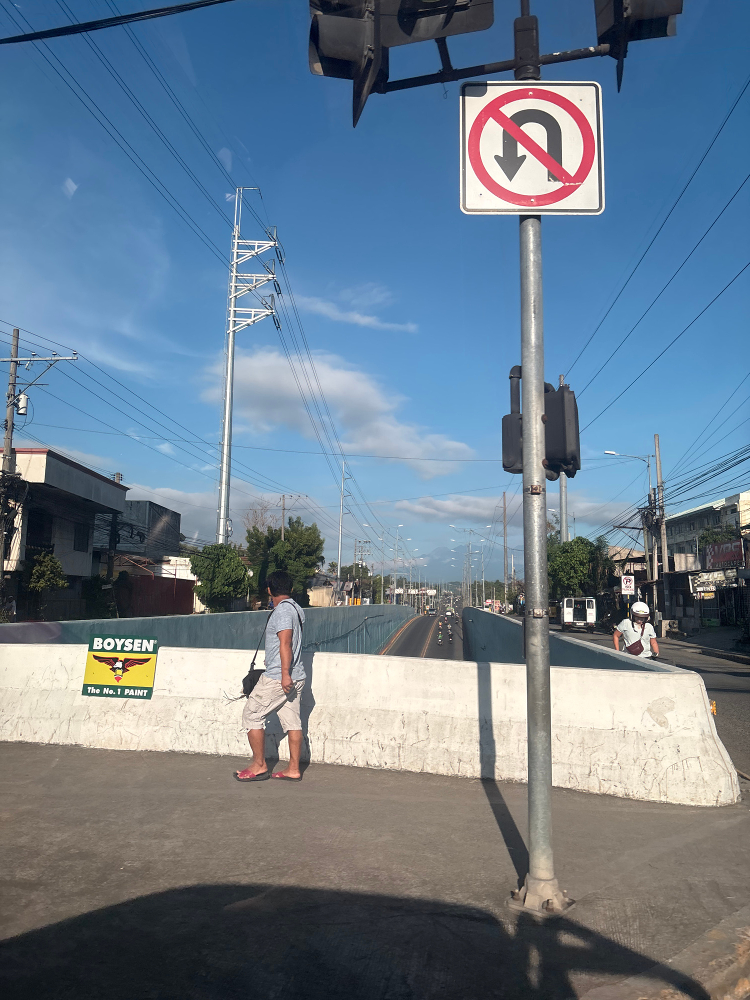The image depicts a street scene in Davao City, Philippines, under a clear blue sky with scattered clouds. In the foreground, a man walks along a concrete pedestrian overpass, wearing a gray t-shirt, beige shorts, and flip-flops. He carries a dark bag over his shoulder. Adjacent to him on the overpass wall is a yellow sign with a black logo and text that reads "BOYSEN" and "The No. 1 PAINT".
A metal pole supporting traffic signals and a "no U-turn" sign stands prominently on the overpass. The sign features a red circle with a diagonal line through a black U-turn arrow pointing left, indicating that U-turns are prohibited.
Beyond the overpass, a road stretches into the distance, with vehicles including cars, motorcycles, and a white van visible. Power lines crisscross the sky, supported by numerous utility poles. Buildings line the sides of the road, some appearing to be commercial establishments. The overall environment suggests an urban setting with ongoing traffic and infrastructure.