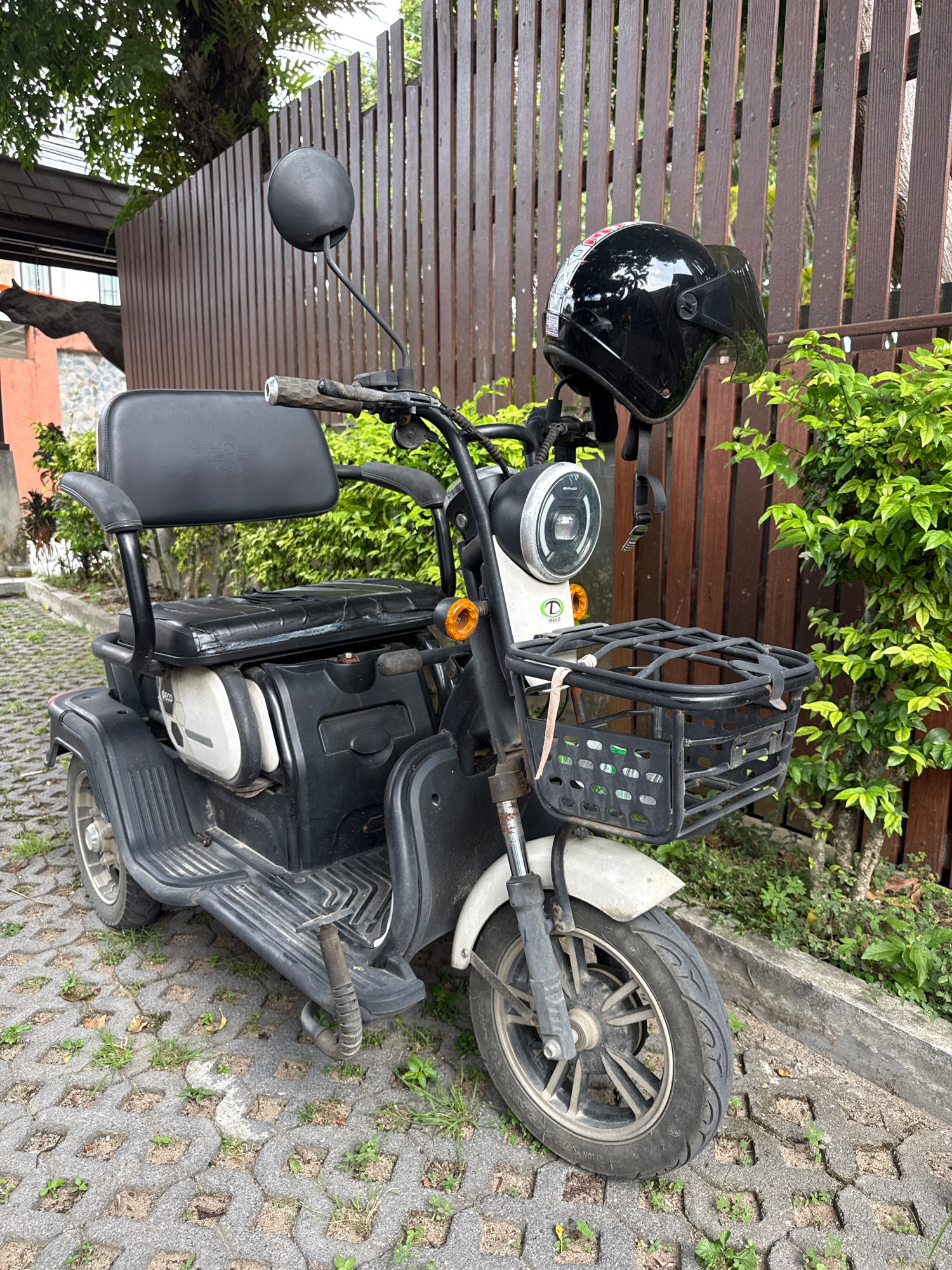 A black three-wheeled electric scooter is parked on a permeable interlocking concrete paver surface in an outdoor environment. The scooter has a black leather seat with armrests and a backrest, a front basket, and a black helmet is hanging from the handlebars. The scooter is positioned in front of a vertical wooden fence and lush green foliage. The scooter's front tire is visible, and the ground shows patches of grass growing through the pavers. The lighting suggests it is daytime. The location is identified as Baan Bo Phut, Thailand.