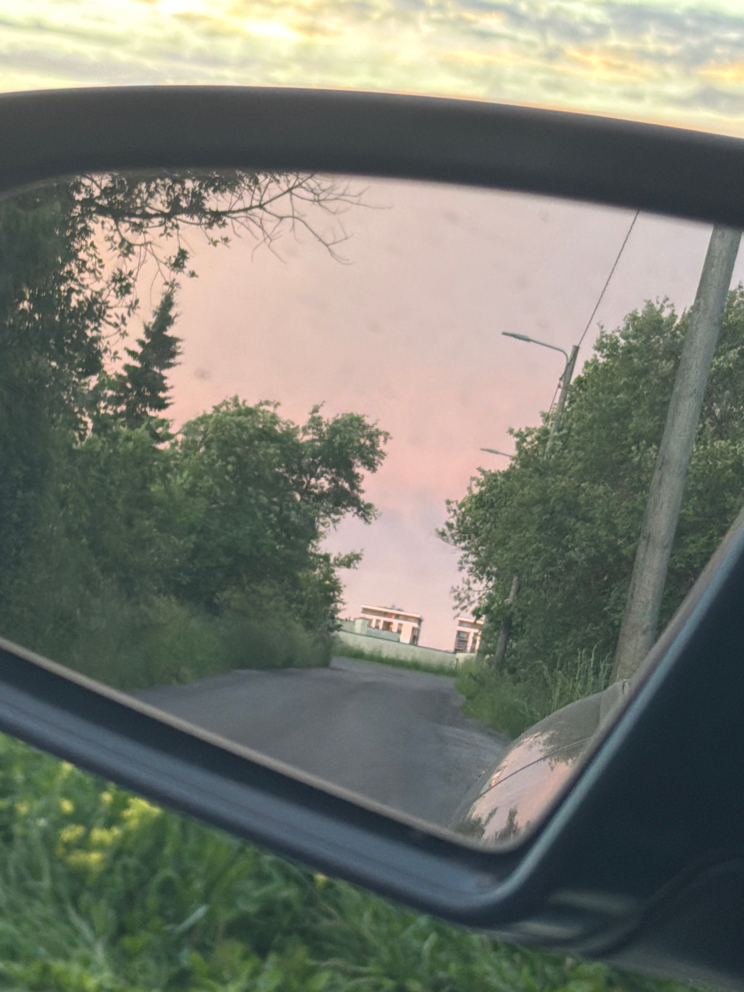 The image shows a reflection in a car's side-view mirror. The reflection captures a rural road with trees lining both sides and a pink and yellow sky with clouds. In the distance, two light-colored buildings are visible. The image is taken during either sunrise or sunset. The location is not identifiable as being in the city of Tallinn, Estonia, or any other specific city.
