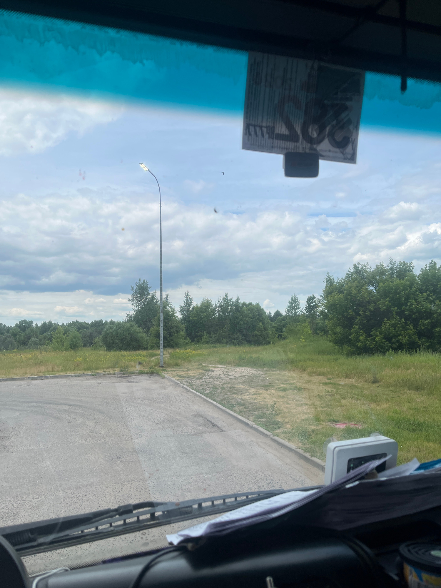 The image shows a view from inside a vehicle, looking out through the windshield. The vehicle is parked on a paved area next to a grassy field with trees in the background. A streetlamp is visible on the left side of the image. There is a sticker attached to the inside of the windshield, partially obscuring the view. The sky is cloudy. The scene appears to be in a rural or semi-rural area. No specific landmarks or identifying features of Chanki, Russia, are visible.