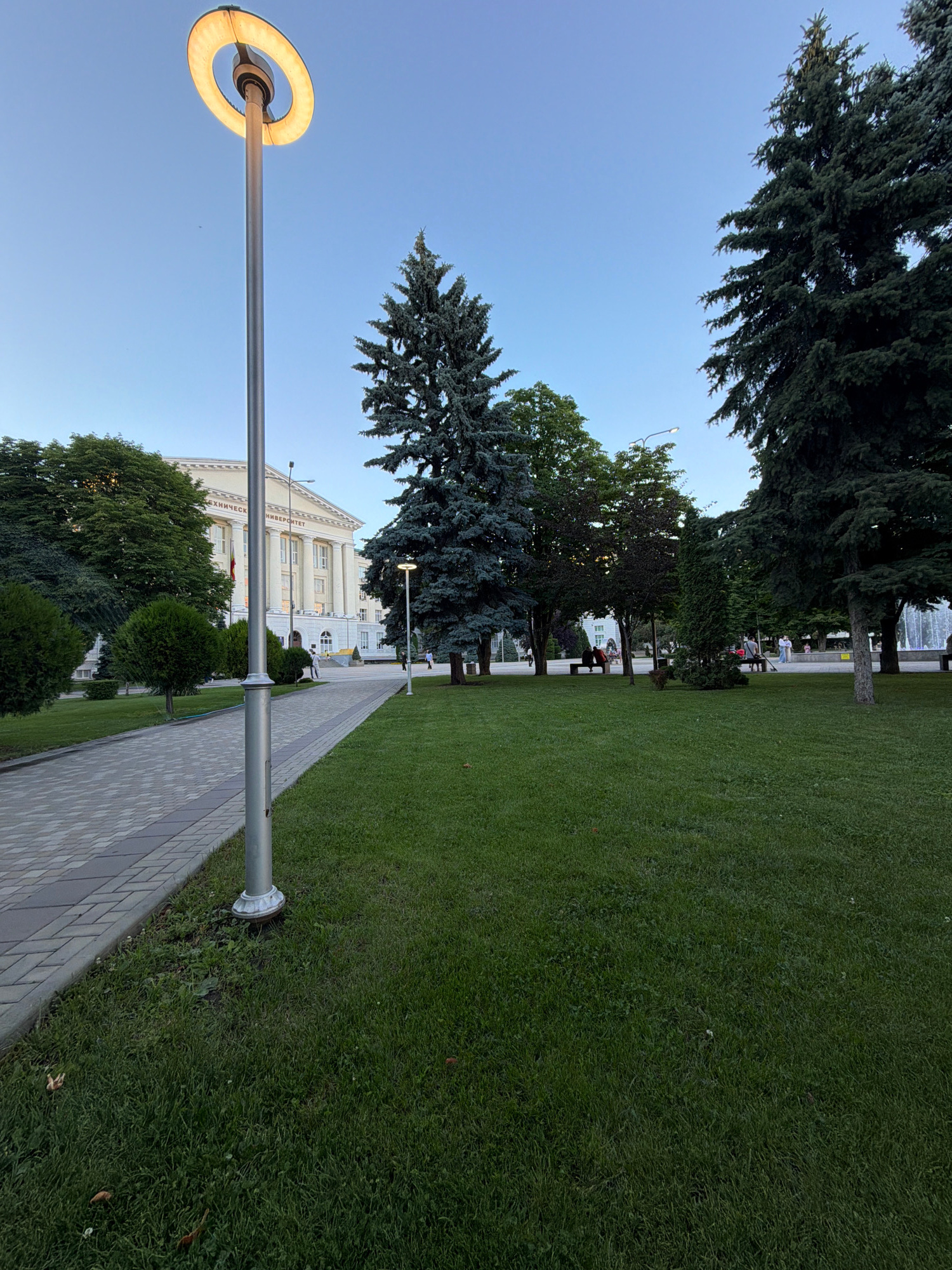 This image depicts a park-like setting with a large, white building in the background. A paved walkway runs along the right side of the image, bordered by a neatly mown green lawn. Several trees, including tall evergreen trees and leafy deciduous trees, are scattered throughout the scene. In the foreground, a modern street lamp with a circular, illuminated head stands beside the walkway.
The building appears to be an institutional or educational building, possibly a university, as suggested by the faint text visible on its facade. The text is in Cyrillic and appears to read "ТЕХНИЧЕСКИЙ УНИВЕРСИТЕТ," which translates to "Technical University."
There are a few people visible in the background, scattered across the lawn and along the walkway. They appear to be casually strolling or sitting. One person is sitting on a bench under a tree. The overall atmosphere suggests a peaceful daytime scene, possibly late afternoon or early evening, given the soft light. The sky is clear and blue, indicating fair weather. The scene is serene and orderly, typical of a public park or campus grounds.