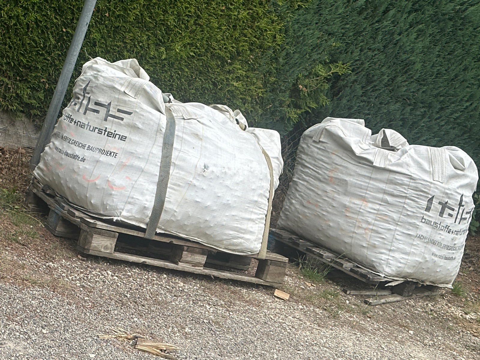 Two large, white, woven bulk bags, likely filled with construction materials such as gravel or sand, are resting on wooden pallets. The bags have black lettering and logos printed on them. The bag on the left has text including "baustoffe+natursteine", "ERFOLGREICHE BAUPROJEKTE", and a website address "raiss-baustoffe.de". The bag on the right has similar text, including "baustoffe+naturs" and "FACHHÄNDLER FÜR ERFOLGREICH ...". The bags are situated outdoors on a gravelly surface, with a dense green hedge in the background. A grey metal pole stands to the left of the left bag. The lighting suggests it is daytime.