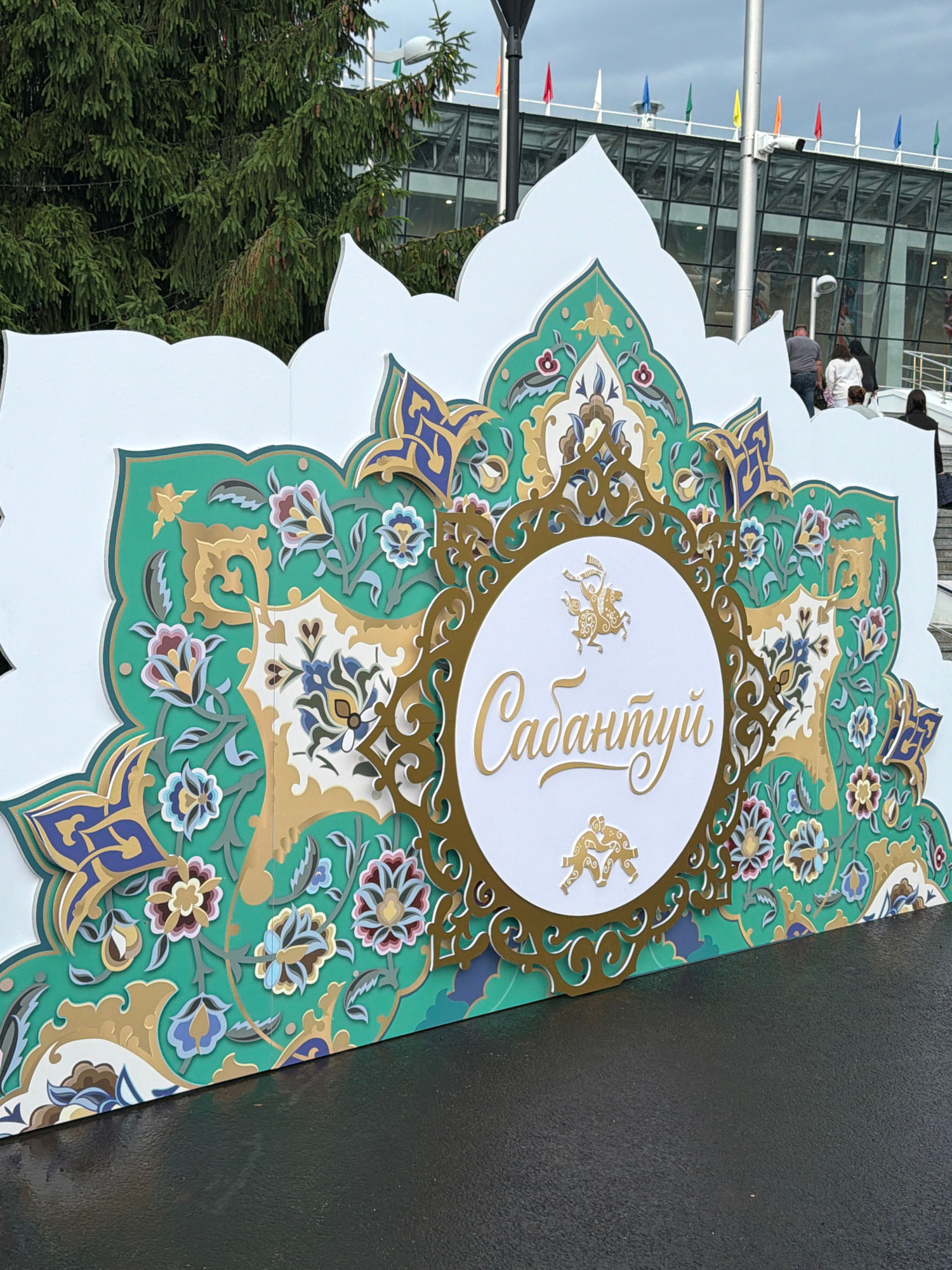 An ornate, multi-layered floral-patterned backdrop with a white circular emblem at its center stands outdoors on a wet, dark asphalt surface. The backdrop is predominantly green with intricate gold, blue, and pink floral and geometric designs. Within the central white circle, stylized golden figures of people appear to be wrestling or performing a dance. Above and below these figures, gold script is visible, with the prominent text reading "Сабантуй." In the background, a modern glass building with a series of colorful flags along its roofline is partially obscured by the backdrop and a large evergreen tree. Several people are walking up a set of stairs towards the building, with their backs to the camera. The sky is overcast.