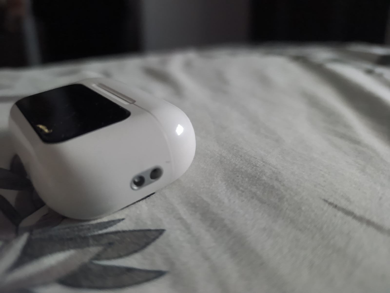A white wireless earbud charging case is positioned on a textured, light-colored surface, possibly a bedspread or blanket with a subtle floral pattern in gray. The case is partially obscuring the foreground of the image. A dark, reflective surface, likely a screen or lid, is visible on the top of the case. Two small, dark circular openings, possibly microphones or sensors, are present on the side of the case, towards the front. The background is out of focus and appears dark. The lighting is soft and diffused, creating a slightly reflective sheen on the case. The scene is set in an indoor environment, with no identifiable urban landmarks.