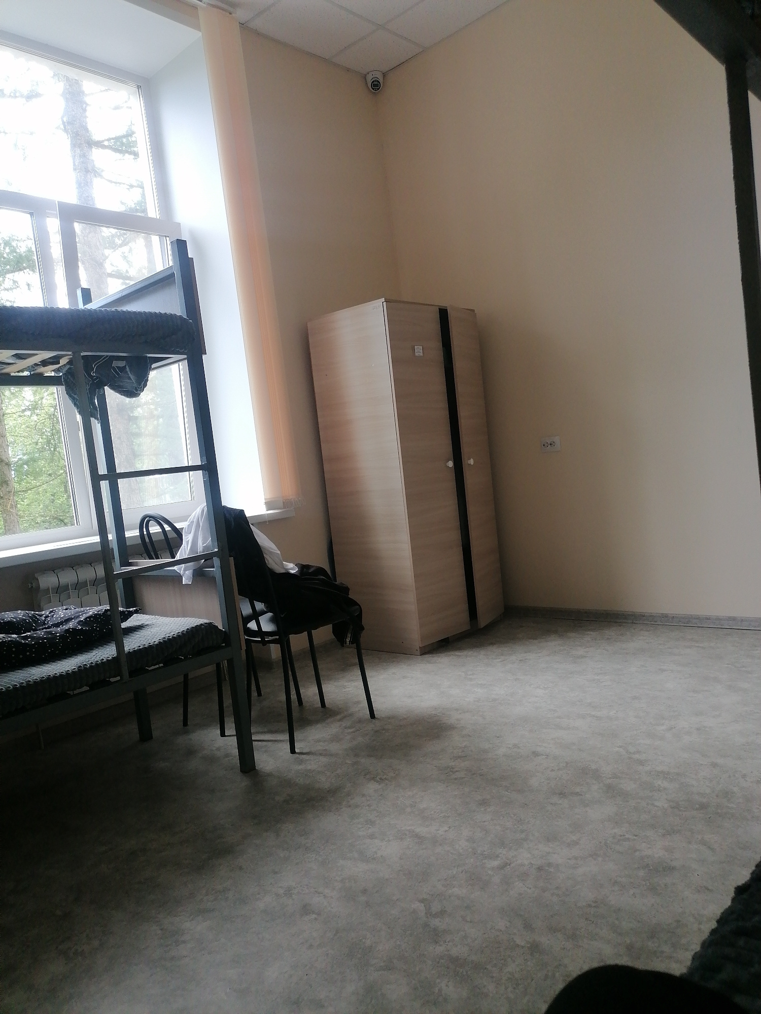 The image depicts a dormitory room. A bunk bed with patterned bedding is visible on the left, with the lower bunk made up and the upper bunk partially covered by a blanket. A metal ladder is attached to the bunk bed. To the right of the bed, a black chair with clothing draped over it, including a white shirt and a black jacket, sits near a window. The window overlooks trees and a bright sky. A light-colored wardrobe with two doors and silver handles stands against the right wall. In the upper corner of the room, a security camera is mounted on the wall. An electrical outlet is visible on the wall near the wardrobe. The floor is a mottled gray, and the walls are painted a light beige.