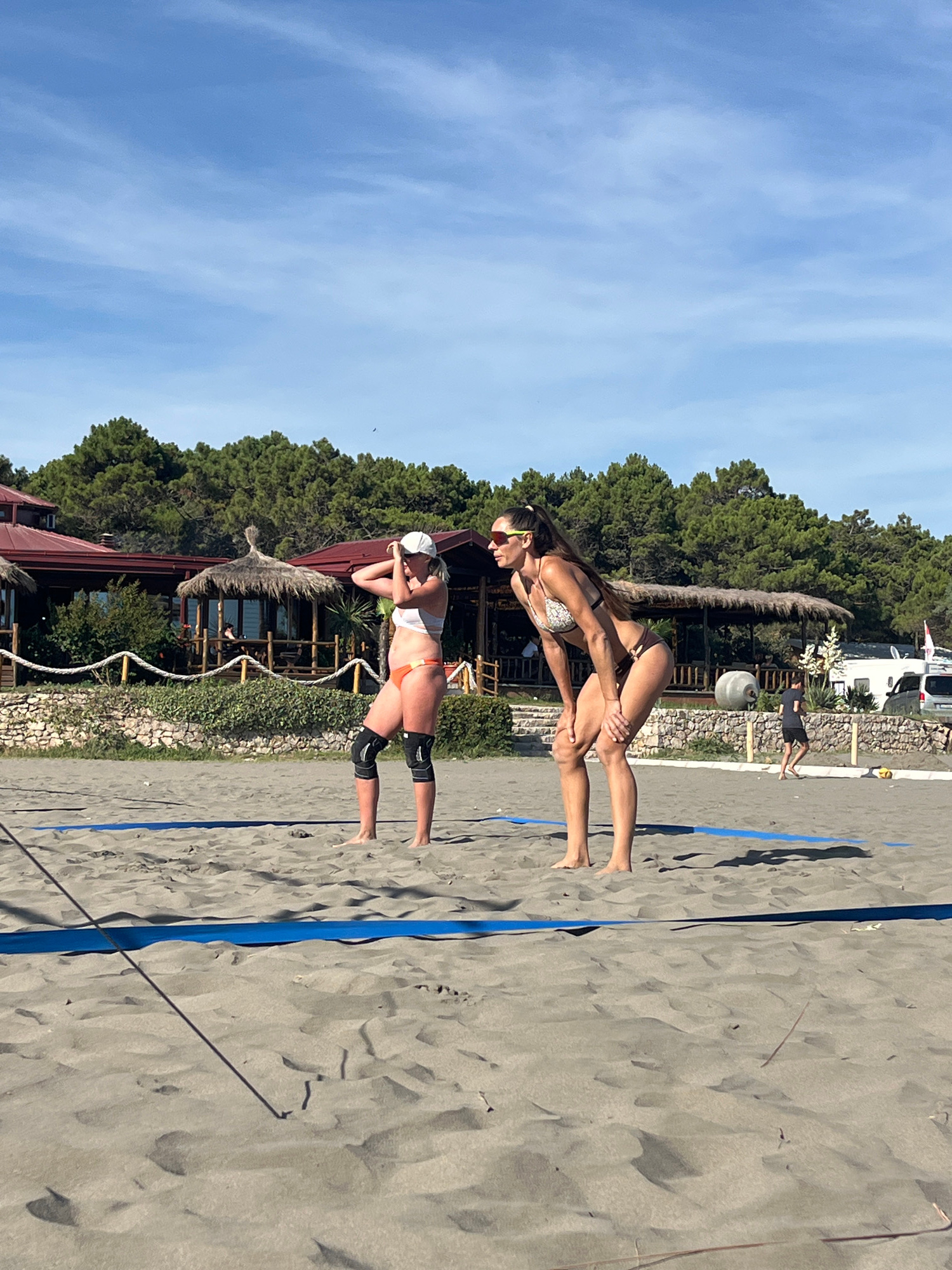 Two women are playing beach volleyball on a sandy court in Pistula, Montenegro. The woman closer to the foreground is wearing an orange bikini and knee pads, in a defensive stance with her arms extended. The second woman, slightly further away and positioned behind and above the first, is wearing a patterned bikini and appears to be in the process of setting or hitting the ball. Her body is contorted in an athletic pose as she reaches forward.
The scene is set on a sunny day, with a clear blue sky and some wispy clouds. Lush green trees form a backdrop behind the volleyball court, and a few rustic buildings with thatched roofs are visible among them. A white camper van is parked in the background, suggesting a holiday or recreational area. The sand on the court is a dark grey, and the blue boundary lines of the volleyball court are clearly visible. The overall impression is one of an active and enjoyable beach activity in a picturesque location. No visible text is present in the image.