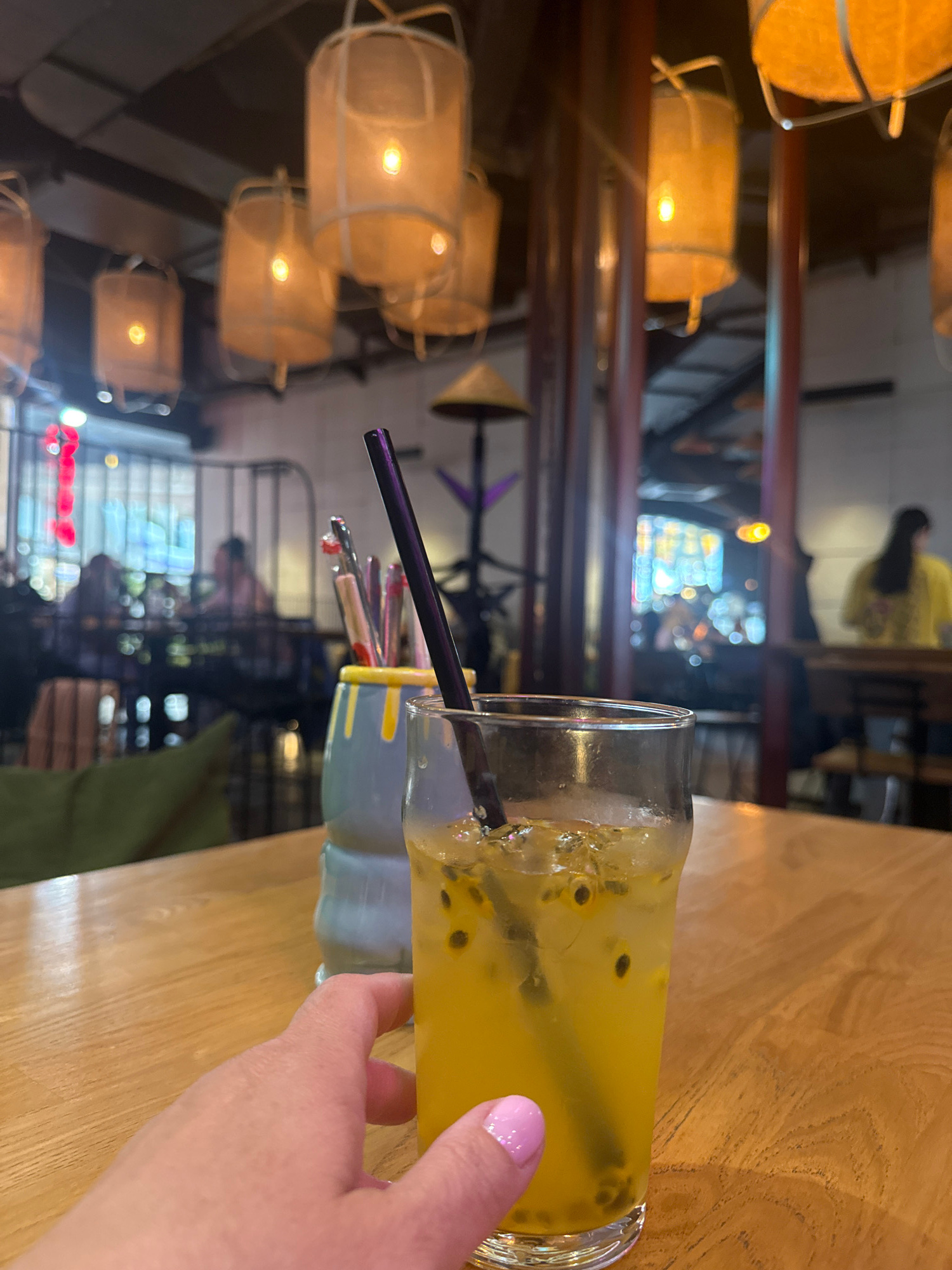A first-person perspective shot shows a person's hand with pink fingernails reaching for a glass of passion fruit juice on a wooden table. The drink is a vibrant yellow, filled with ice and passion fruit seeds, and has a black straw. Behind the drink, a ceramic holder with straws and utensils is visible. The background depicts a dimly lit restaurant interior with several ornate, illuminated lanterns hanging from the ceiling. In the blurred background, patrons are seated at tables, and a person in a yellow shirt is visible. The scene appears to be set in the city of Irkutsk, Russia, potentially at a restaurant such as "Salsa Bar" known for its Asian-inspired decor and lighting.