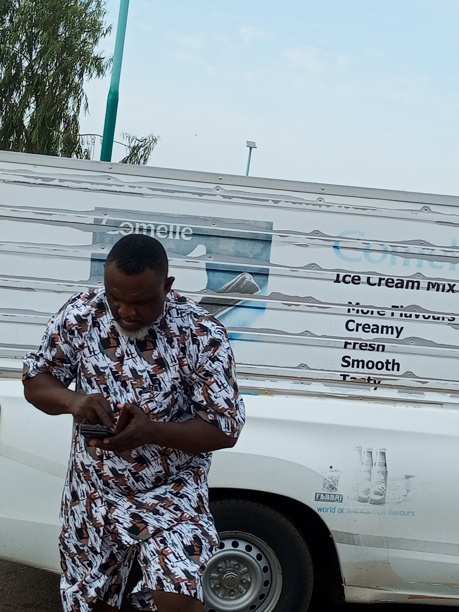 A man with a beard and wearing a patterned shirt is looking down at a small black object he is holding in his hands. He is standing next to a white vehicle that has "Ice Cream Mix" written on the side. Below this, the words "More Flavours," "Creamy," "Fresh," "Smooth," and "Tasty" are also visible. Further down on the side of the vehicle, there is a logo that reads "FABBRI" with the tagline "world of authentic flavours." The vehicle appears to be a refrigerated truck or van, suggested by the corrugated metal siding and the visible tire and wheel.
In the background, a tall green pole stands against a bright, overcast sky. Trees with green foliage are visible behind the pole. The overall scene suggests an outdoor setting, possibly during the daytime, given the brightness of the sky. The man's focus on the object in his hands might indicate he is using a phone or another small device. The setting appears to be an urban or semi-urban area, given the presence of the vehicle and the man.