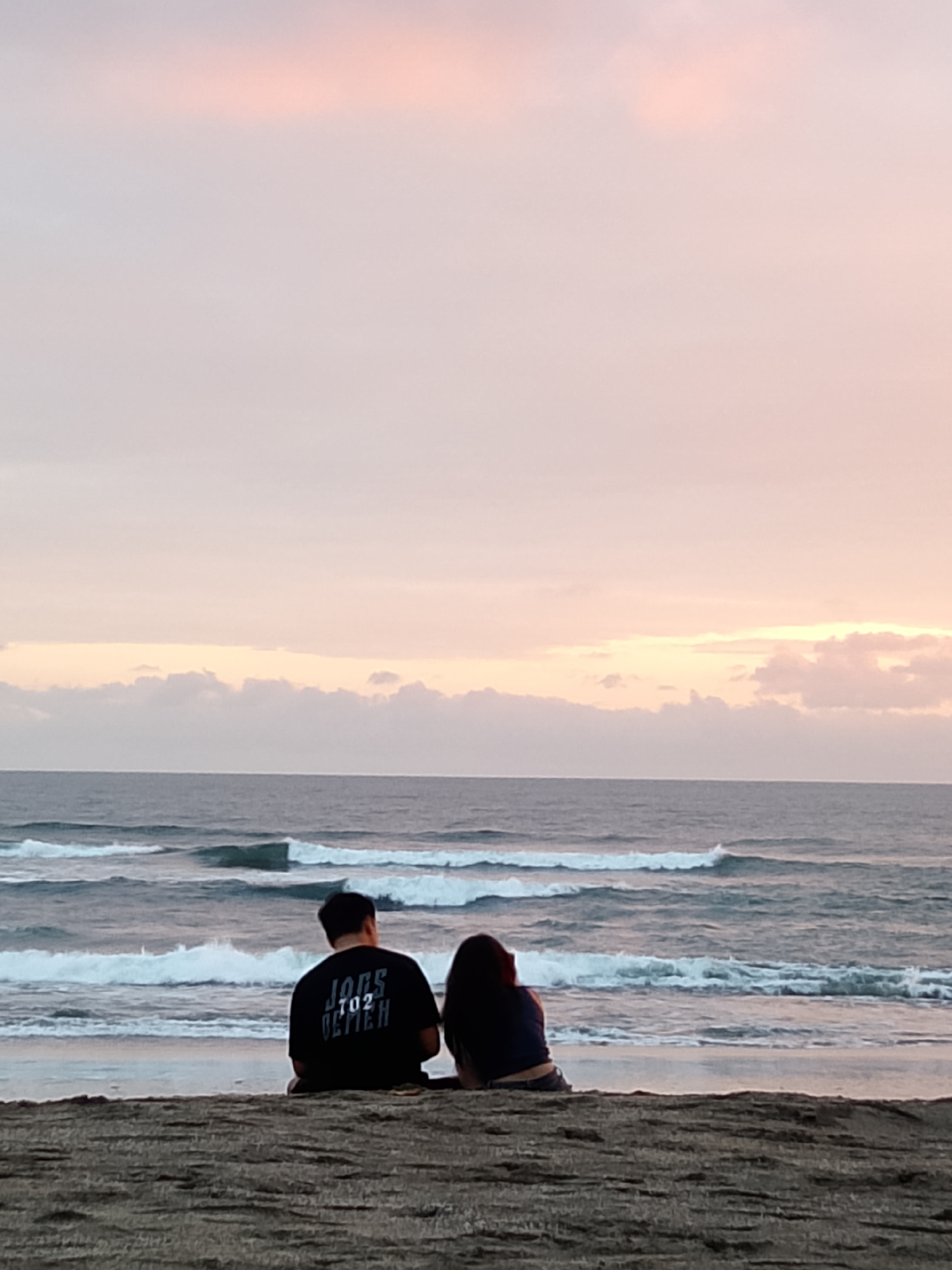 An outdoor, ground-level, medium shot captures two individuals from behind, seated on a sandy beach. The person on the left wears a black t-shirt with white lettering that reads "JUGS 702 ULYMPH." The person on the right has dark, shoulder-length hair and wears a dark top and denim shorts. They are positioned facing a tranquil ocean with visible waves, illuminated by the soft, pastel hues of a sunset sky. The foreground consists of dark, coarse sand. The scene suggests a peaceful moment at a beach, potentially located in the city of Kediri, Indonesia, during twilight.