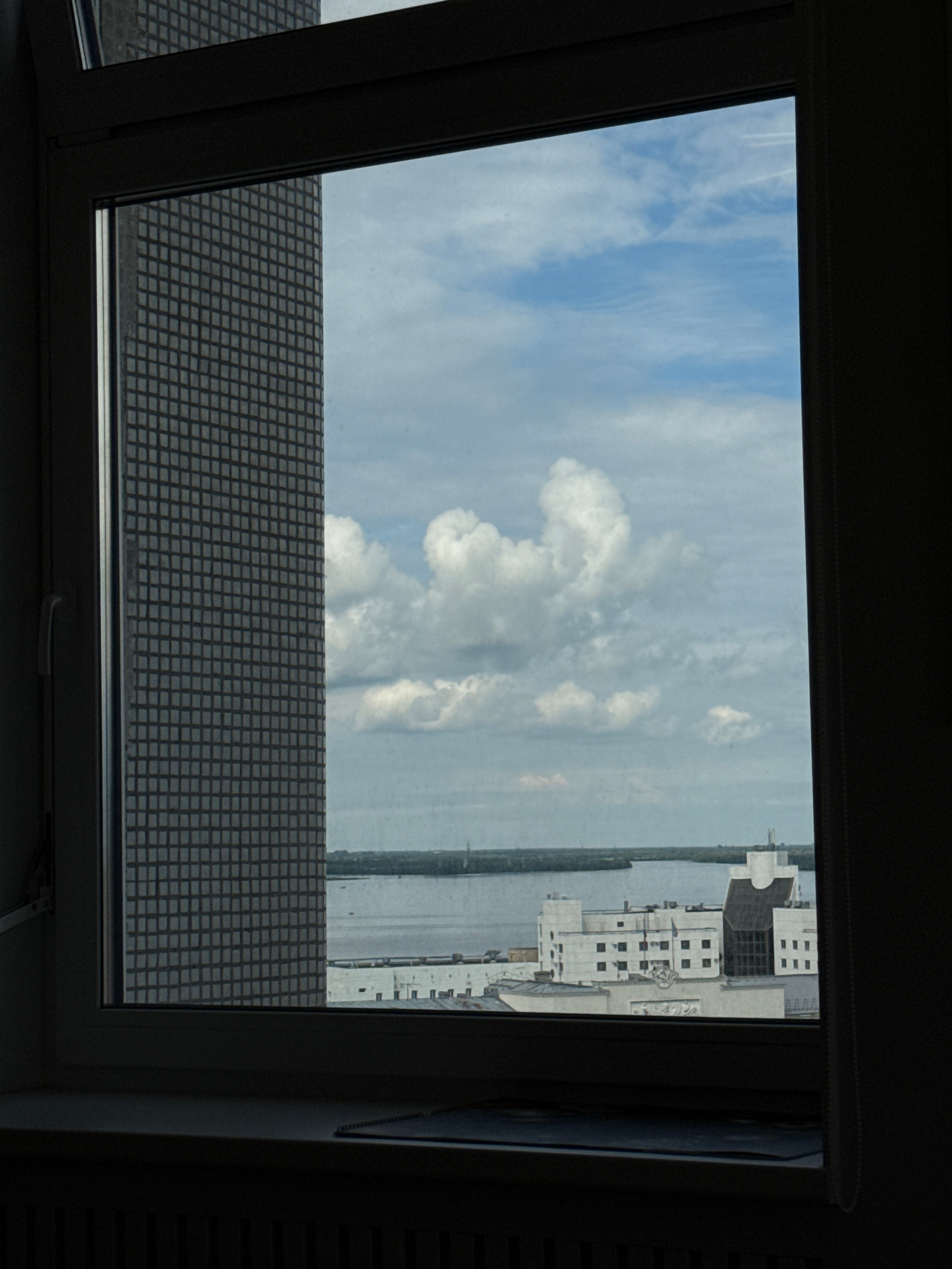 The image shows a view through a window in a high-rise building, likely in Arkhangelsk, Russia. To the left, a portion of a gray, grid-patterned building is visible, obscuring the view. The majority of the frame is filled with a sky dominated by white, cumulus clouds against a pale blue backdrop. In the distance, across a body of water, a cluster of white, modern buildings with dark windows can be seen. One building has a distinct, curved roofline and a large, central glass facade. The water appears calm, with a few small boats visible on its surface. The foreground shows the dark interior of the room with the window frame and a dark curtain on the right side. A dark windowsill with a radiator below is also visible. The overall scene suggests an urban environment with a view of a waterfront.