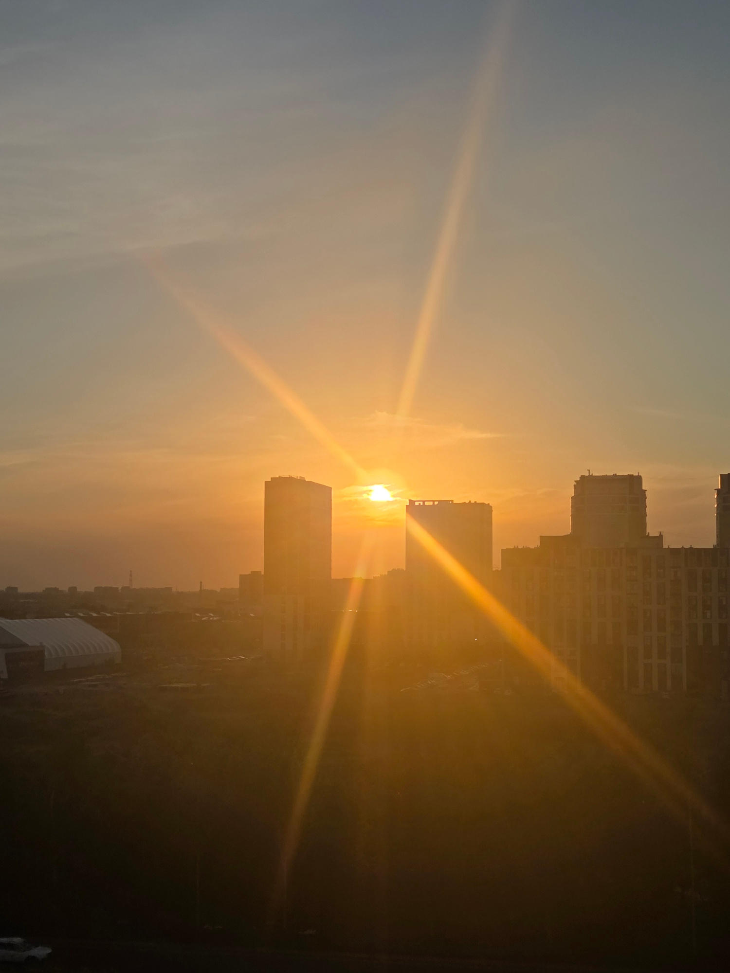 The image depicts a cityscape at sunset, oriented vertically. The sun is low on the horizon, casting a warm, golden glow and creating strong light rays that stretch across the scene. Silhouetted against the bright sky are several tall, modern buildings, their outlines sharp and defined. The buildings appear to be apartment blocks or office buildings. In the foreground, more buildings and some foliage are visible, largely obscured by the intense sunset light, making them appear dark and indistinct. The sky transitions from a pale blue on the left to a vibrant orange and yellow closer to the sun. The overall impression is one of a peaceful end to the day in an urban environment. The time of day is clearly sunset due to the position of the sun and the warm, low-angled light. The weather appears clear, with no clouds visible to obstruct the sun. There are no visible people or text in the image. The location is identified as Karaganda, Kazakhstan.