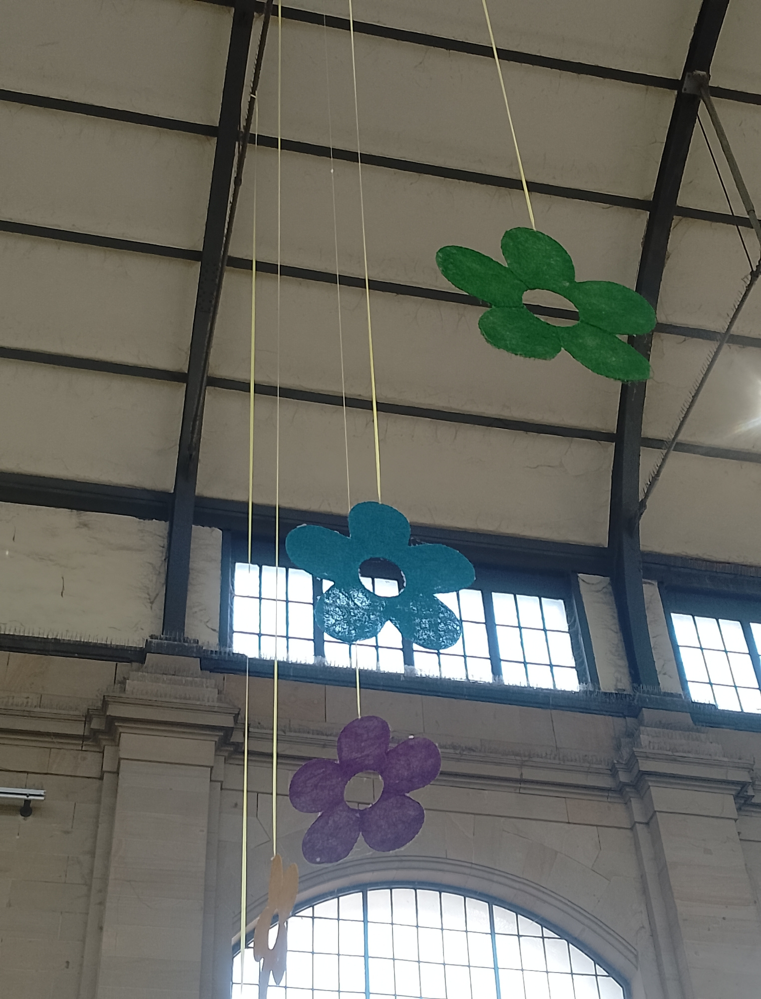 The image shows a ceiling structure with hanging decorations. The ceiling is made of a light-colored material and supported by dark metal beams. There are several strings hanging down, each with a cutout flower decoration. The flowers are in different colors: green, blue, purple, and yellow. The green flower is at the top, followed by the blue, then the purple, and finally the yellow one at the bottom. The background shows large windows with a grid pattern, suggesting an indoor space with ample natural light. The overall impression is of a decorative display, possibly for an event or as a permanent fixture in a building. There is no visible text in the image.