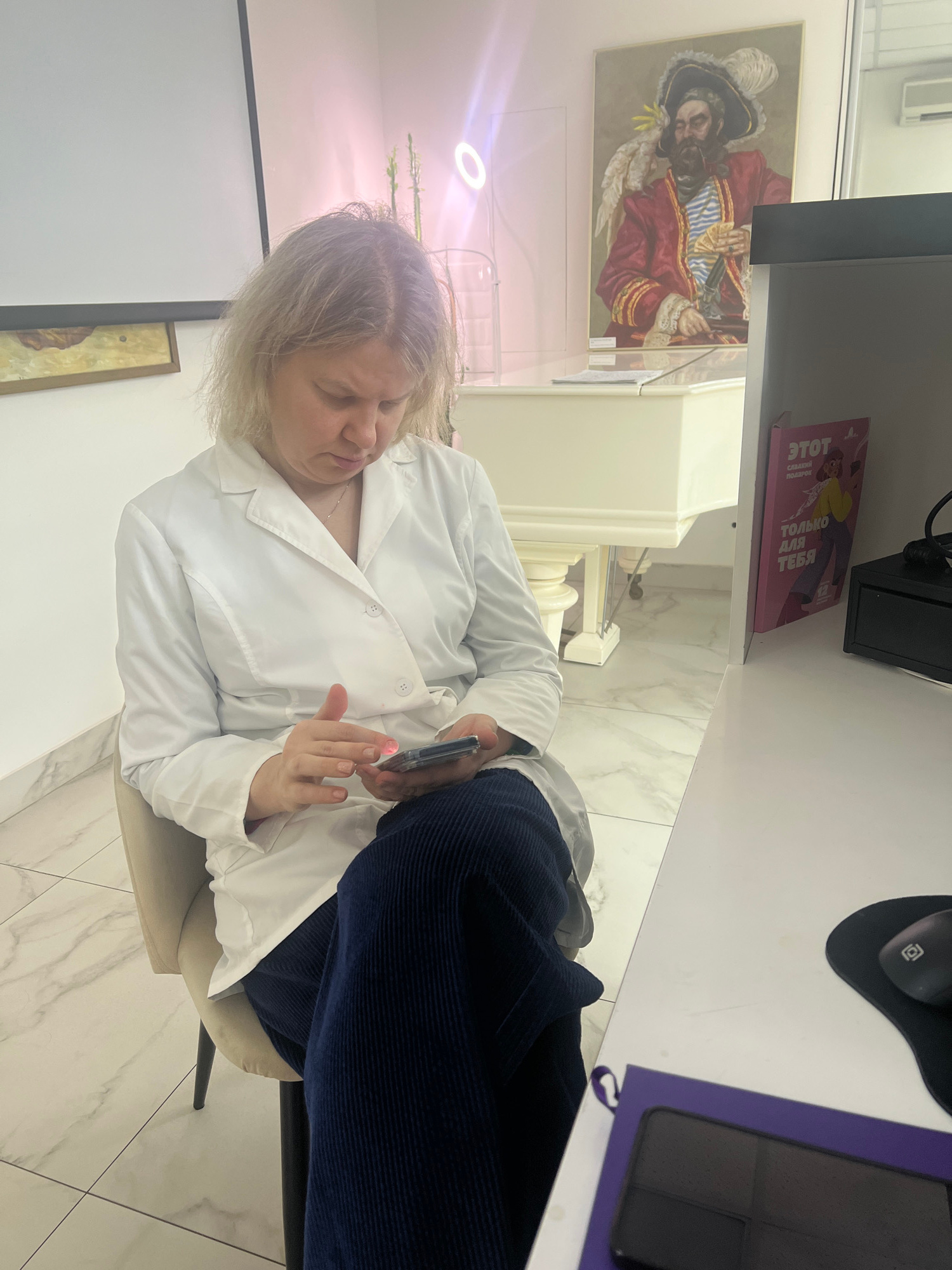 The media file shows a woman in a white lab coat sitting at a desk. She appears to be holding a small, dark object in her right hand, possibly a phone or a pen, and is looking down at it. Her left hand is visible, with fingernails painted pink. The woman has short, light-colored hair and her eyes are closed or she is looking downward intently. She is wearing a dark blue, corduroy garment on her legs.
The setting appears to be an office or medical facility with modern marble-patterned flooring and light-colored walls. Behind the woman, a white piece of furniture resembling a cabinet or a desk is visible. To the left, a framed painting of a man in a red, ornate coat and hat, reminiscent of a pirate or historical figure, is hanging on the wall. On the desk, to the right of the woman, a dark computer mouse on a mousepad and a purple-bound object, possibly a tablet or book, are present. A pink book with Cyrillic text is also visible on a shelf above the painting. The lighting suggests it is daytime, and the indoor setting indicates no specific weather conditions. The overall mood seems focused and calm.
The visible text on the pink book includes "ЭТОТ" and "ТОЛЬКО ДЛЯ ТЕБЯ" in Cyrillic script, which translates to "THIS" and "ONLY FOR YOU". The painting on the wall also has some text on a small label below it, which is illegible. The scene is located in Moscow, Russia.