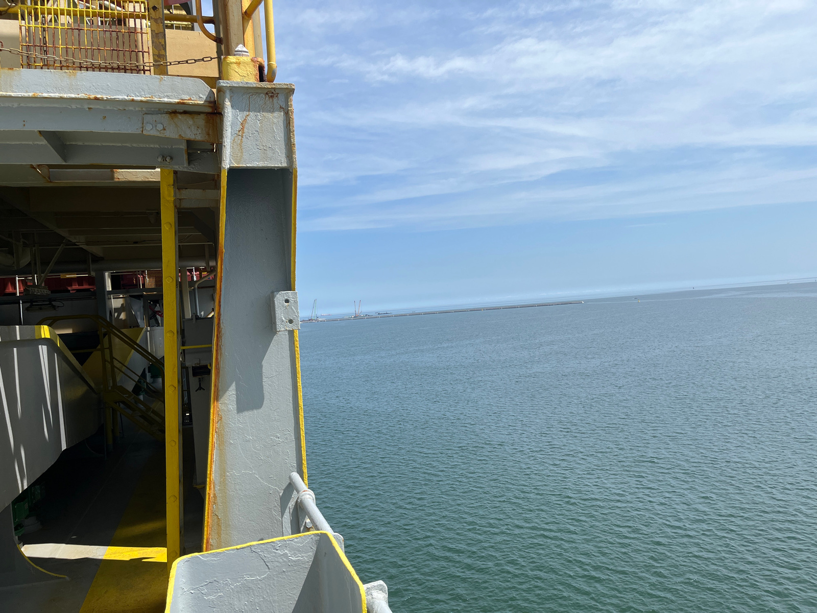 This image captures a view from a ferry or ship, looking out towards the sea. The foreground is dominated by the metallic structure of the vessel, painted in shades of grey and yellow, with visible signs of rust, suggesting an industrial or utilitarian setting. Part of a stairway is visible within the ship's interior, indicating movement or access to different levels.
The main subject is the vast expanse of the Baltic Sea, stretching out to the horizon under a partly cloudy sky. The water is a muted blue-green, with gentle ripples on its surface. In the far distance, a long, man-made structure, likely a breakwater or pier, stretches across the water. Several cranes and other structures are visible along this distant feature, suggesting ongoing construction or port activity. The horizon is a clear line separating the sea from the sky.
The time of day appears to be late morning or early afternoon, given the brightness of the light and the angle of the shadows cast by the ship's structure. The weather seems pleasant, with scattered white clouds against a pale blue sky.
The location is identified as Gdańsk, Poland. While there are no explicit text elements visible in the image, the presence of port infrastructure and the sea strongly aligns with this coastal city. The overall mood is one of openness and journey, with the vastness of the sea and sky creating a sense of calm and perhaps anticipation.