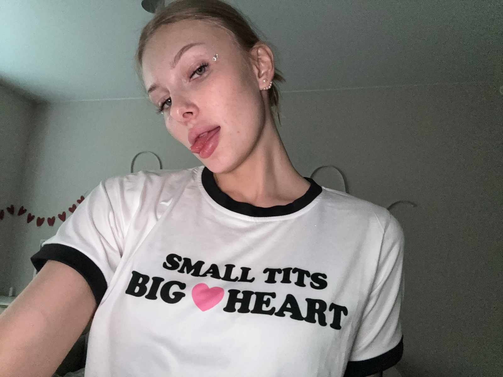 A young woman with blonde hair pulled back is pictured from the chest up, looking at the camera. She has glitter applied to her face near her eye and on her cheekbone, and is sticking her tongue out. She is wearing a white t-shirt with black trim around the collar and sleeves. The t-shirt has black text that reads "SMALL TITS" on the top line, and "BIG HEART" with a pink heart symbol in place of the word "BIG" on the bottom line. The background is dimly lit and appears to be an indoor space, possibly a bedroom, with a wall visible behind her. There are heart-shaped decorations hanging on the wall to the left of the woman. No specific location in the city of Perm, Russia can be determined from the image.