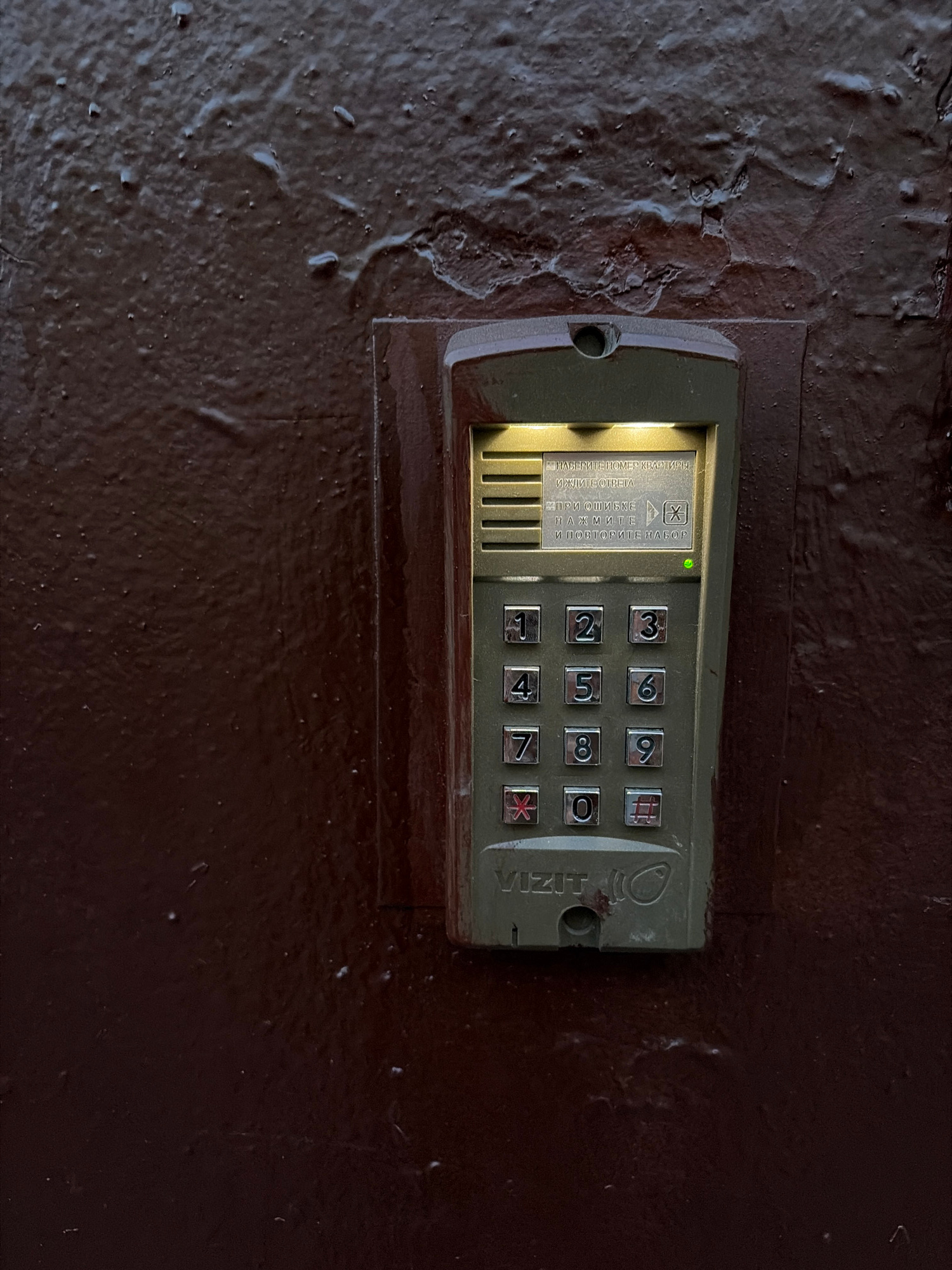 A close-up, high-angle shot captures a metallic intercom system mounted on a textured, dark brown wall. The intercom unit itself is rectangular with rounded corners and is a muted green color, showing signs of wear and minor damage. A keypad with illuminated numbers from 0 to 9, a star symbol, and a hash symbol is centrally located on the unit. Above the keypad, a small screen displays text in Cyrillic, and a green indicator light is illuminated. The screen text translates to: "ENTER APARTMENT NUMBER AND PRESS ANSWER. IF YOU MAKE A MISTAKE, PRESS AND REDIAL." Below the keypad, the "VIZIT" brand name is visible. The wall surrounding the intercom is uneven and appears to be painted metal, with a glossy finish that reflects light in places. Small droplets and textured imperfections are visible on the wall's surface. The scene is indoors or in a sheltered outdoor area, indicated by the controlled lighting and the absence of natural elements like sky or foliage.
