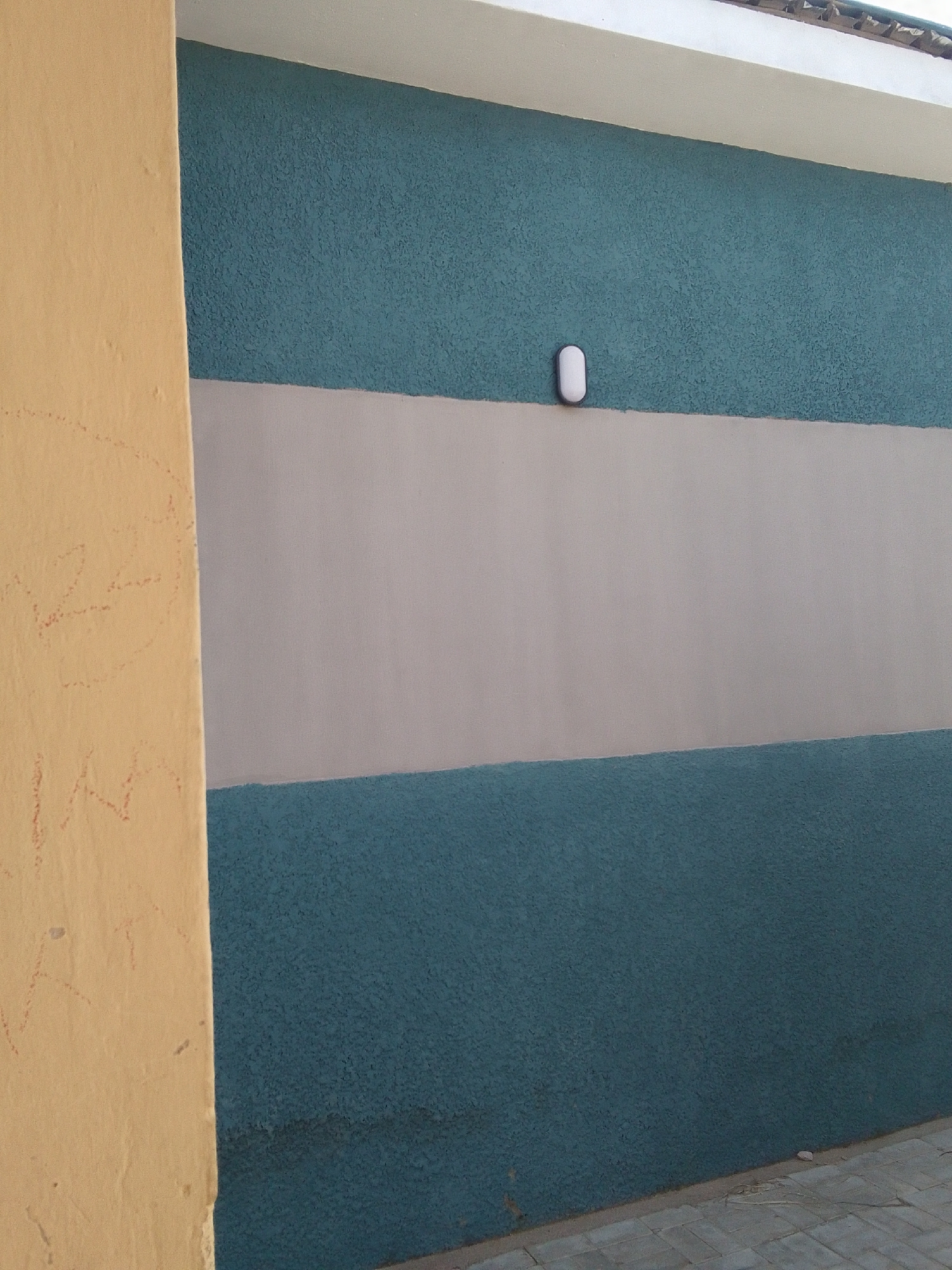 The image shows a close-up view of a textured, horizontally striped wall in an outdoor setting. The wall is divided into three sections: a top section of teal, a middle section of light grey, and a bottom section of teal. A white, oval-shaped light fixture is mounted on the teal section, just above the grey stripe. To the left of the wall, a narrow, beige column or pillar is visible, with faint red markings that appear to be graffiti or drawings. The ground in front of the wall consists of light-colored paving stones. The overall scene is austere and lacks any human presence or discernible activity. The architectural style suggests a modern, possibly commercial or residential building.