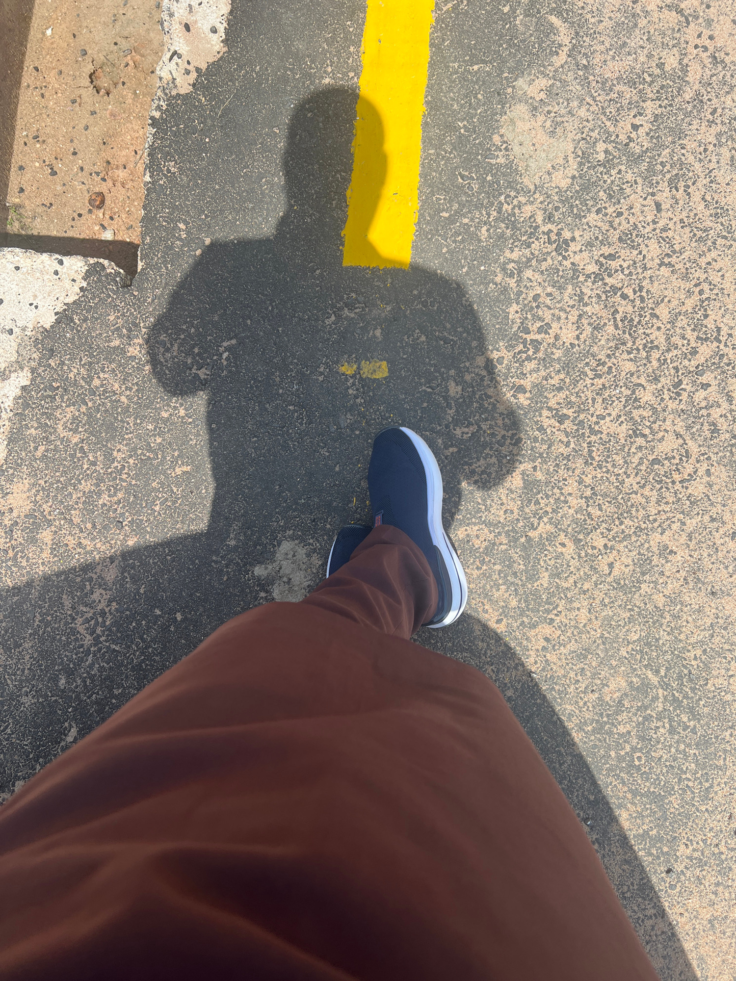 The image captures a person's lower leg and foot, shod in a dark blue sneaker with a white sole and trim, against a textured asphalt surface. The person is wearing brown trousers that are partially visible. A bright yellow line runs horizontally across the asphalt, and a significant shadow is cast by the person, suggesting it is daytime. The asphalt itself appears worn and weathered, with visible cracks and embedded stones. A small section of what looks like a concrete curb or border is visible in the bottom left corner. The lighting suggests a sunny day, likely outdoors. The overall scene is simple and focuses on the textures of the ground and the person's attire.